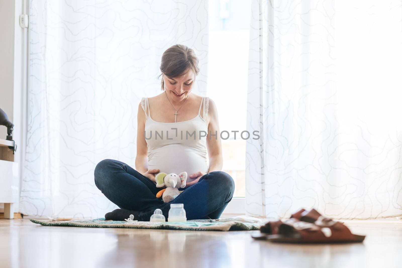 Happy pregnant mother is sitting on the floor, hugging her tummy by Daxenbichler