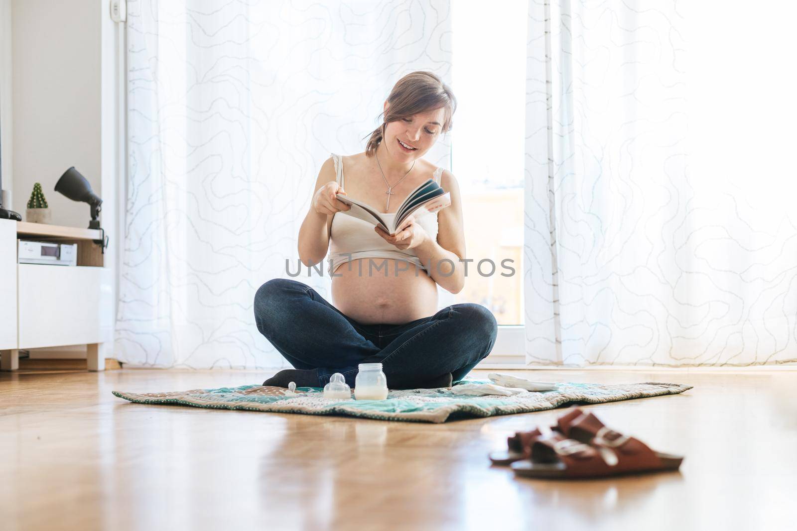 Reading about pregnancy: Happy pregnant mother is sitting on the floor, reading a book by Daxenbichler