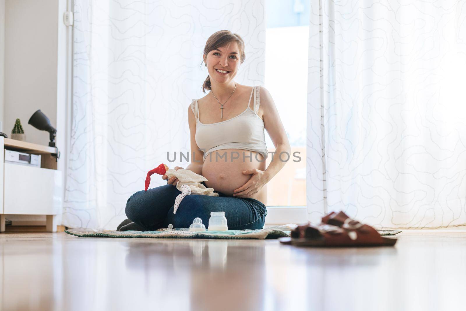 Happy Caucasian pregnant mother is sitting on the floor, touching her tummy, blue jeans