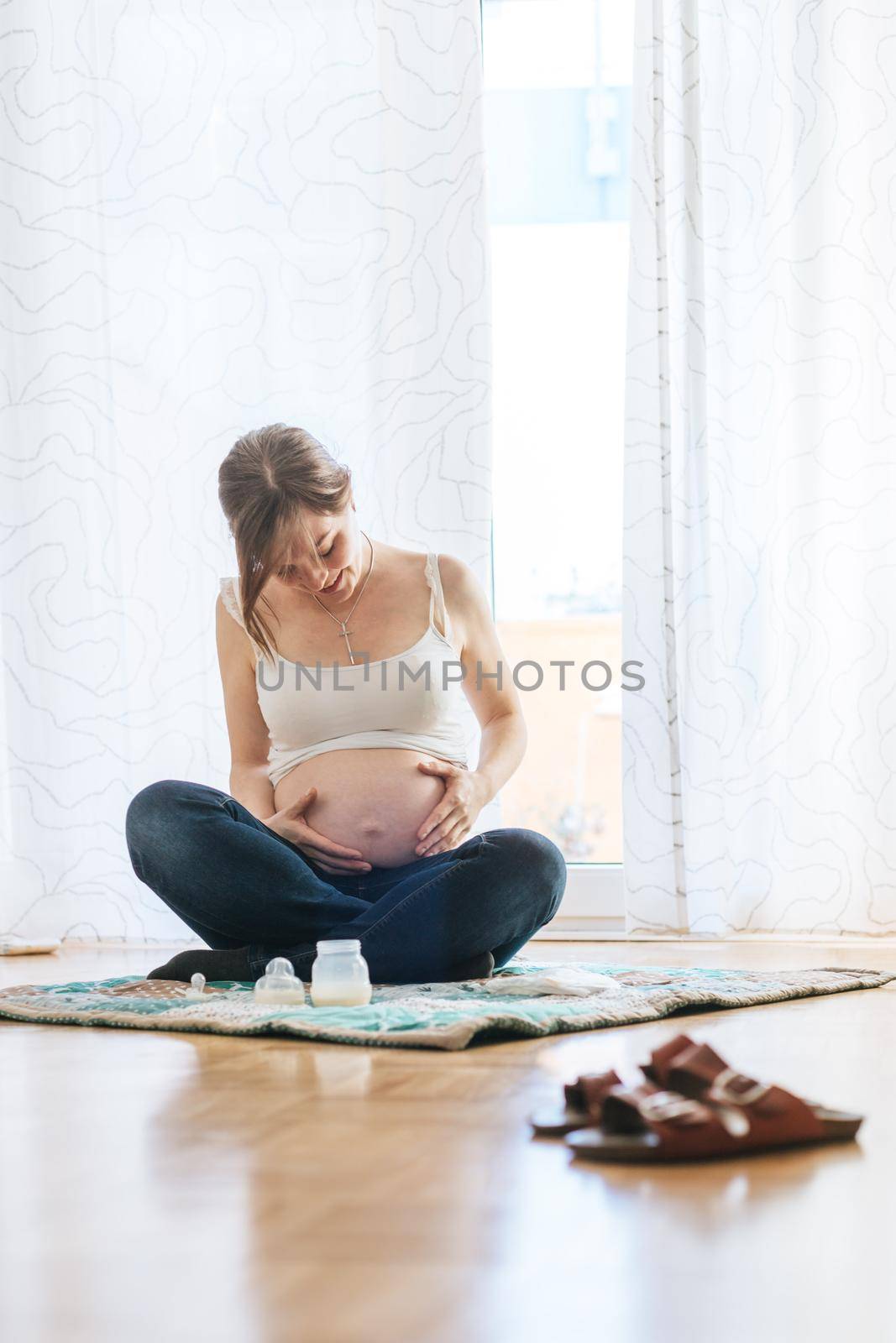 Happy Caucasian pregnant mother is sitting on the floor, touching her tummy, blue jeans