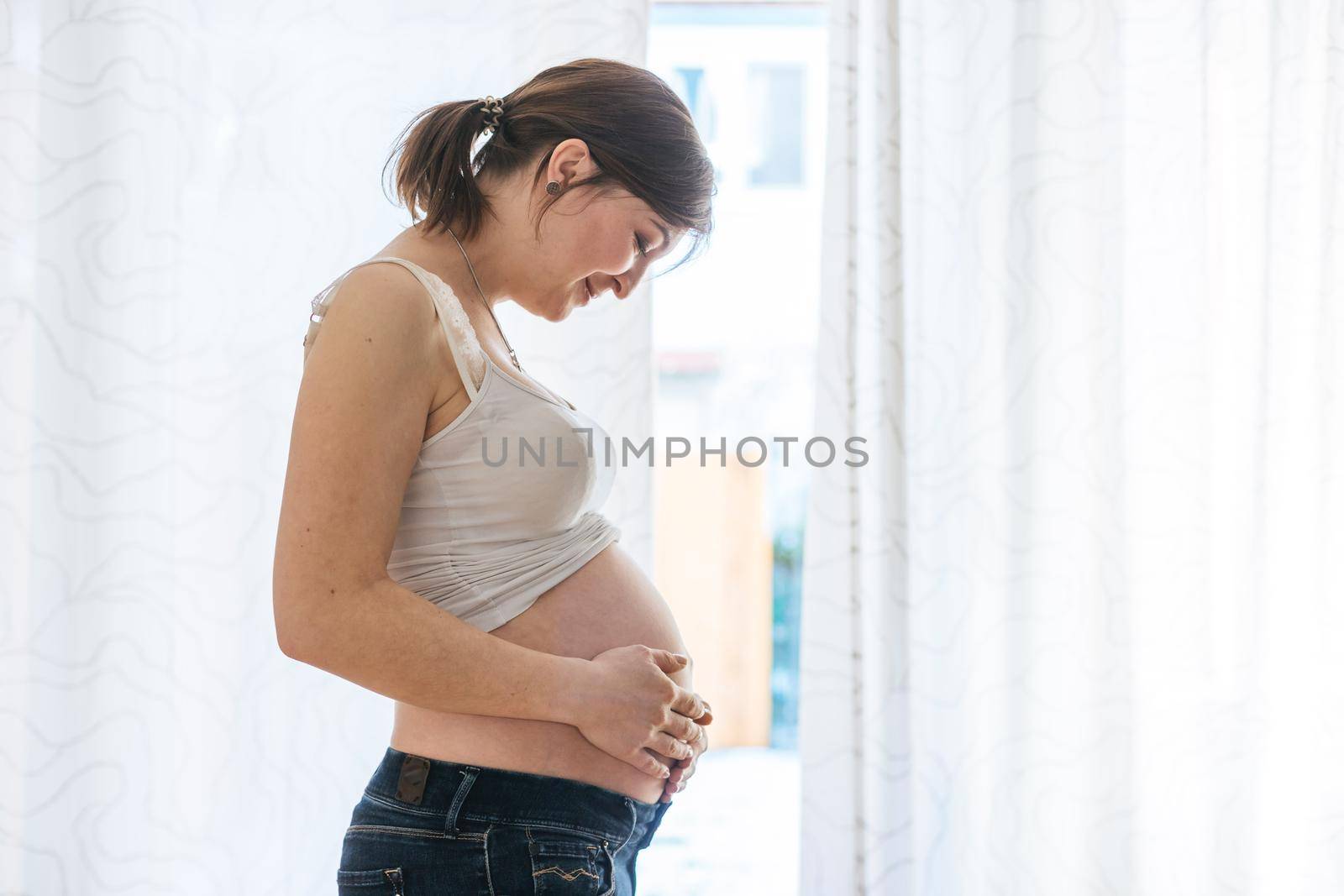 Happy Caucasian pregnant mother touching her naked tummy, blue jeans, close up