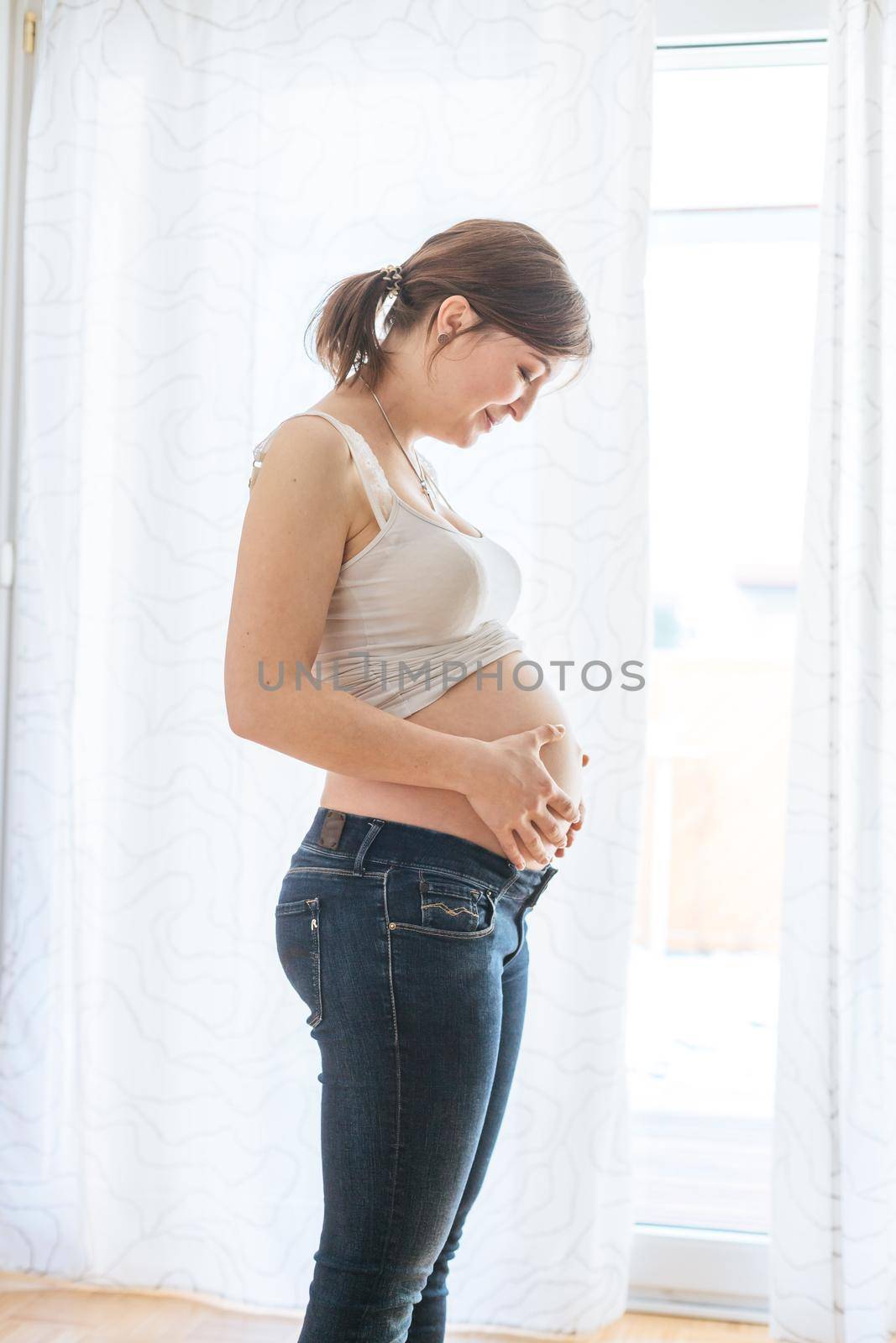 Happy Caucasian pregnant mother touching her naked tummy, blue jeans, close up