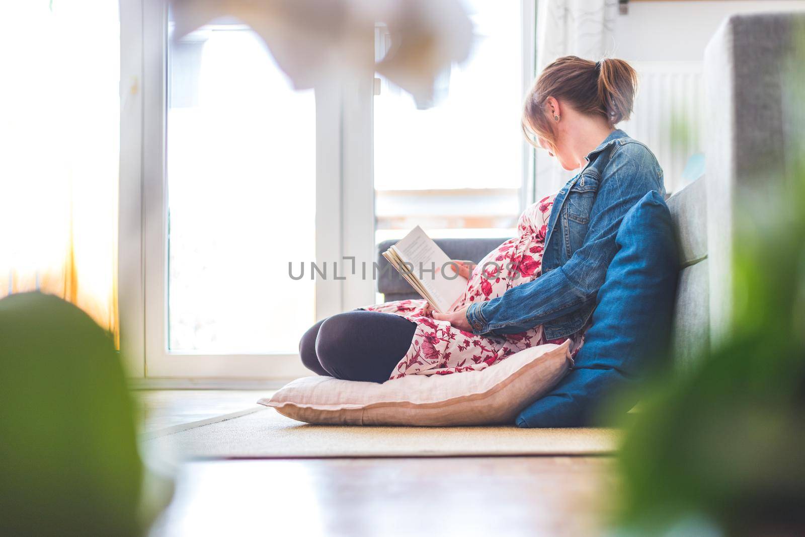 Preparing for pregnancy: Pregnant Caucasian mother is sitting on the floor, reading a book for about babies by Daxenbichler