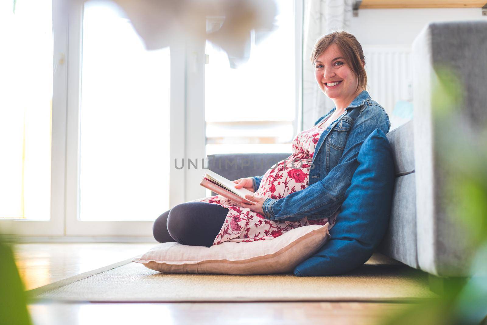 Preparing for pregnancy: Pregnant Caucasian mother is sitting on the floor, reading a book for about babies by Daxenbichler