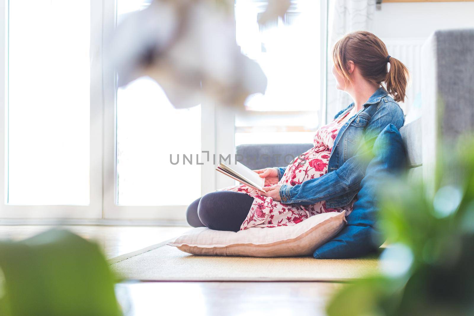 Preparing for pregnancy: Pregnant Caucasian mother is sitting on the floor, reading a book for about babies by Daxenbichler