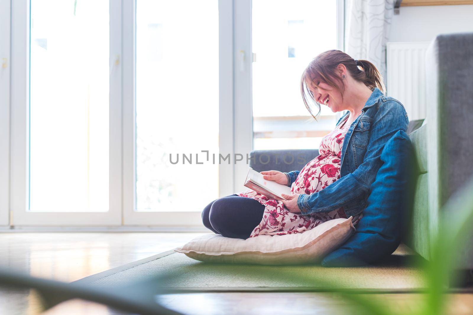 Pregnant caucasian happy mother is sitting on the floor, touching her tummy, floral dress