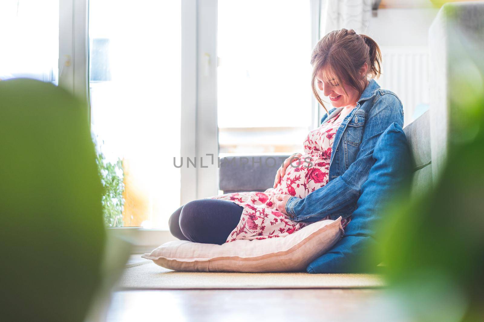 Pregnant caucasian happy mother is sitting on the floor, touching her tummy, floral dress