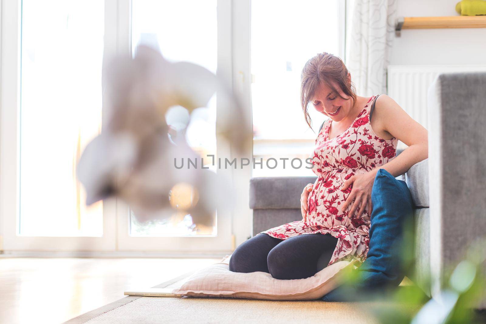 Pregnant caucasian happy mother is sitting on the floor, touching her tummy, floral dress