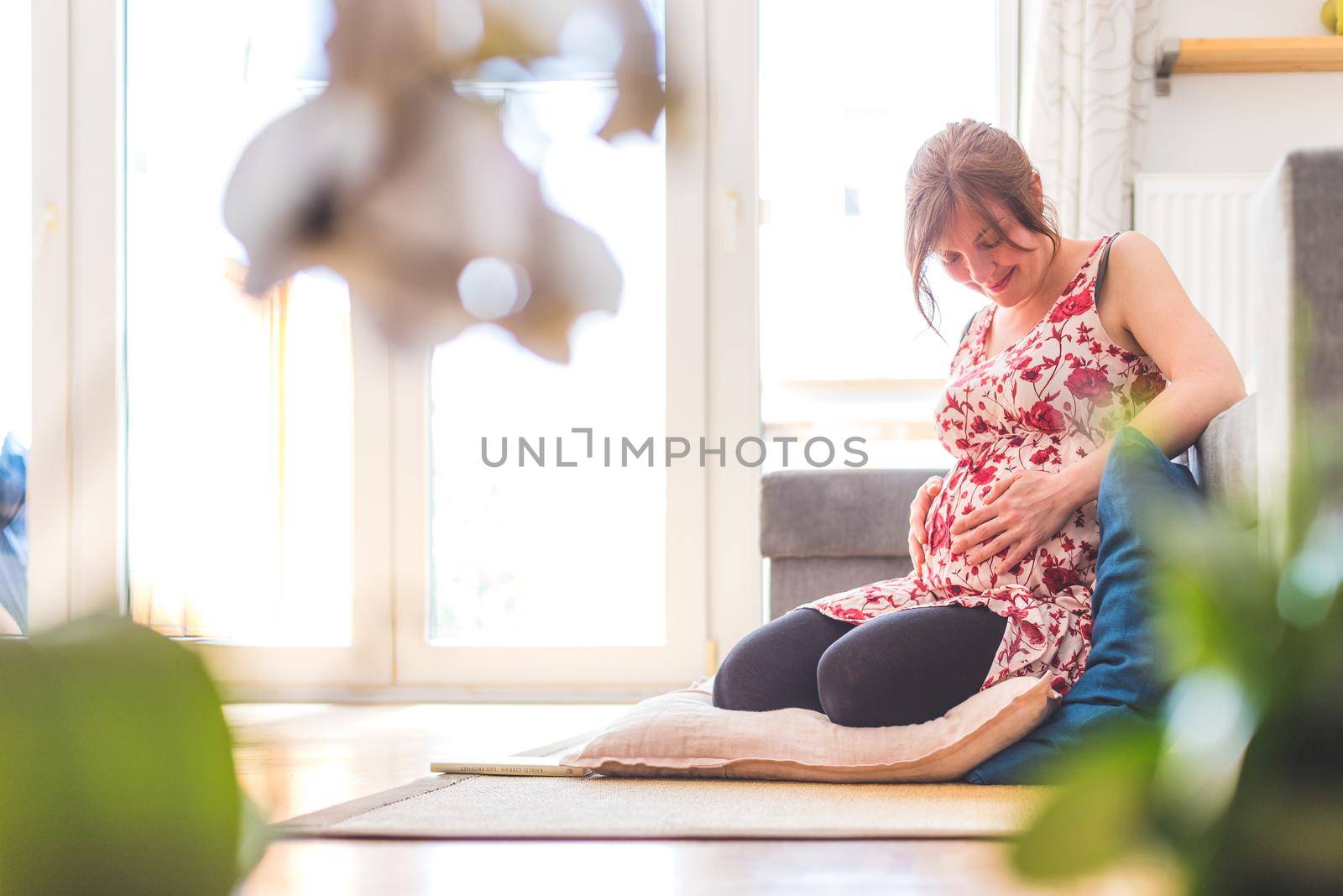 Pregnant caucasian happy mother is sitting on the floor, touching her tummy, floral dress