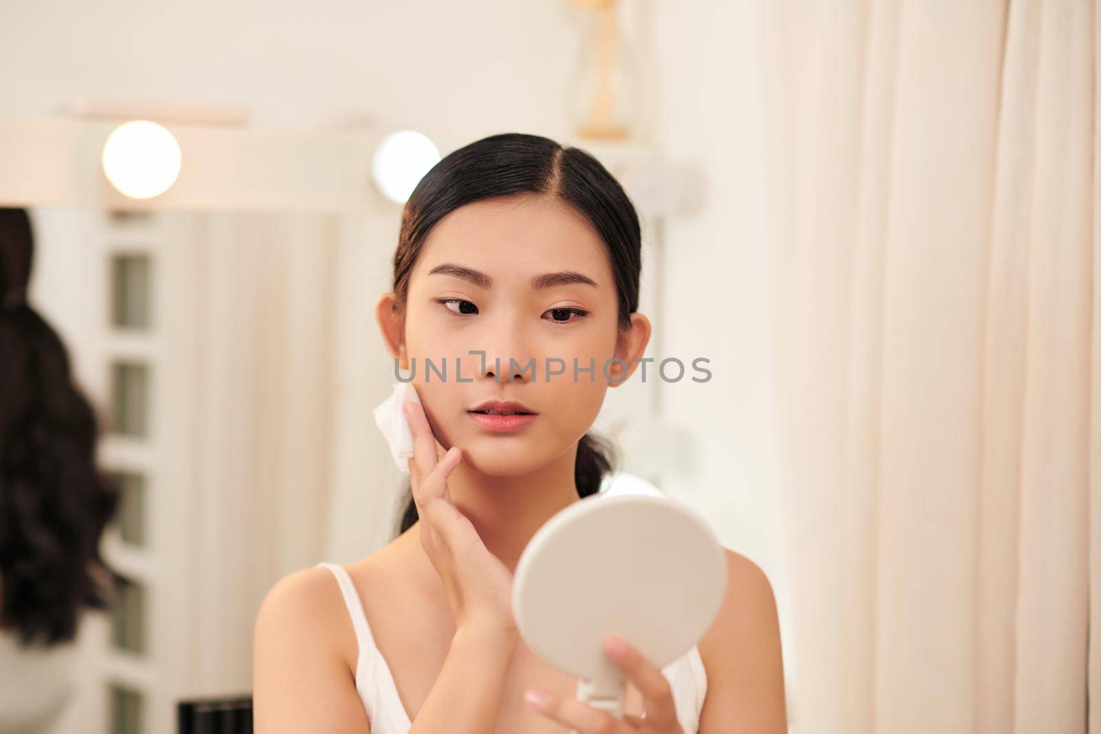 Young cute woman remove makeup , cleaning face with cotton pads looking in mirror by makidotvn