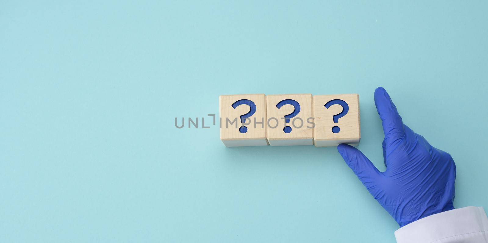 a hand in a blue medical glove holds cubes with question marks on a blue background. The concept of unknown, riddle, unanswered questions, copy space