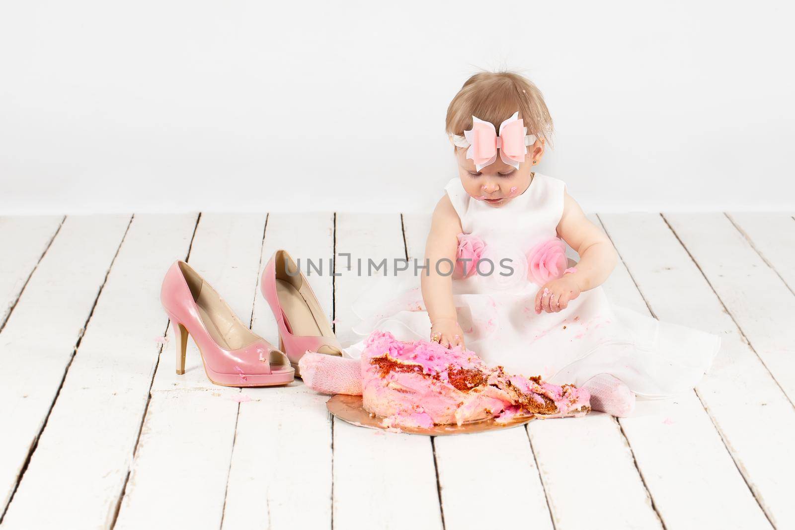Little funny year-old girl with a festive cake, smeared with cream, is sitting on the floor.