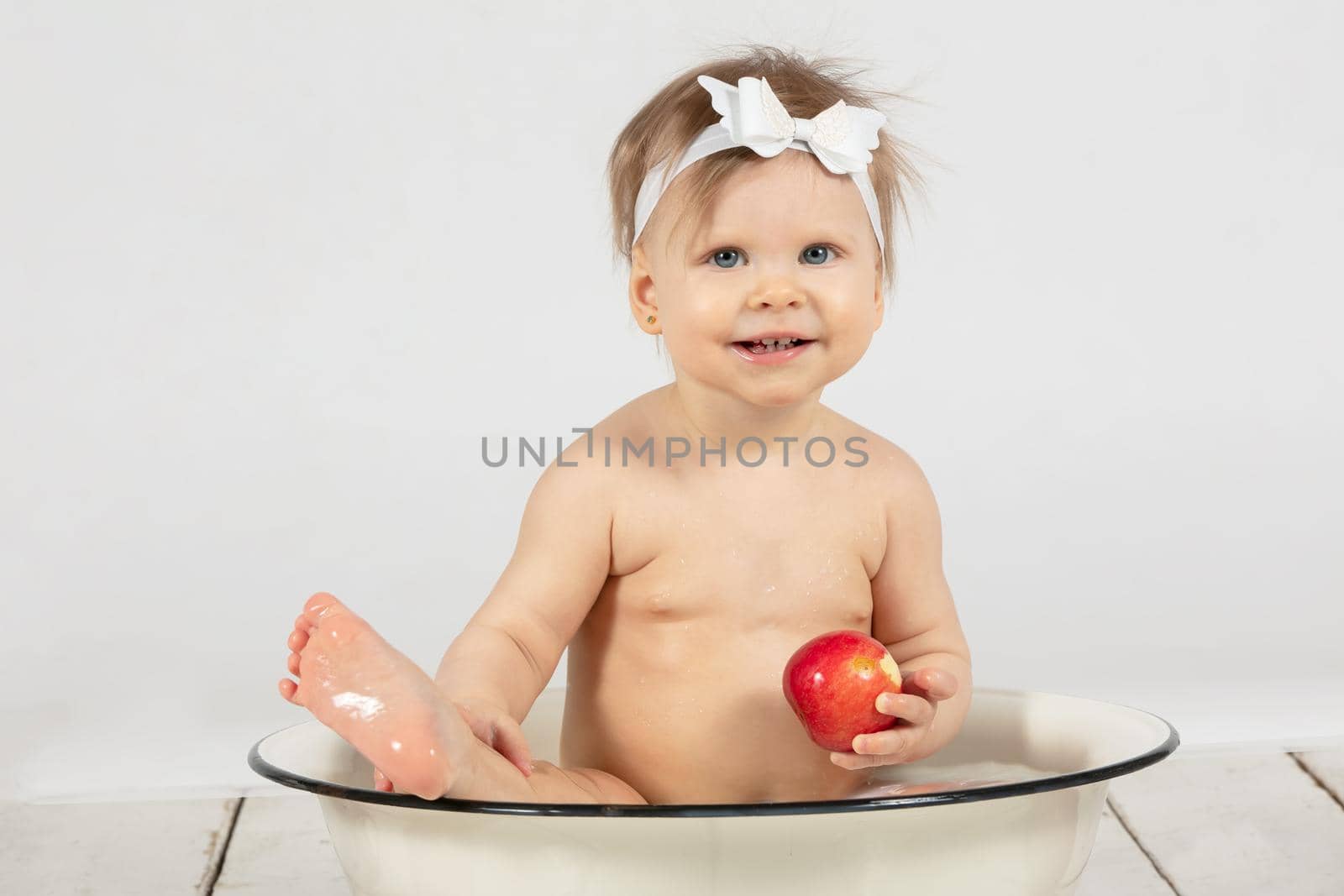 Beautiful baby bathes in a basin and eats a red apple. Little girl in one year old. Funny child.