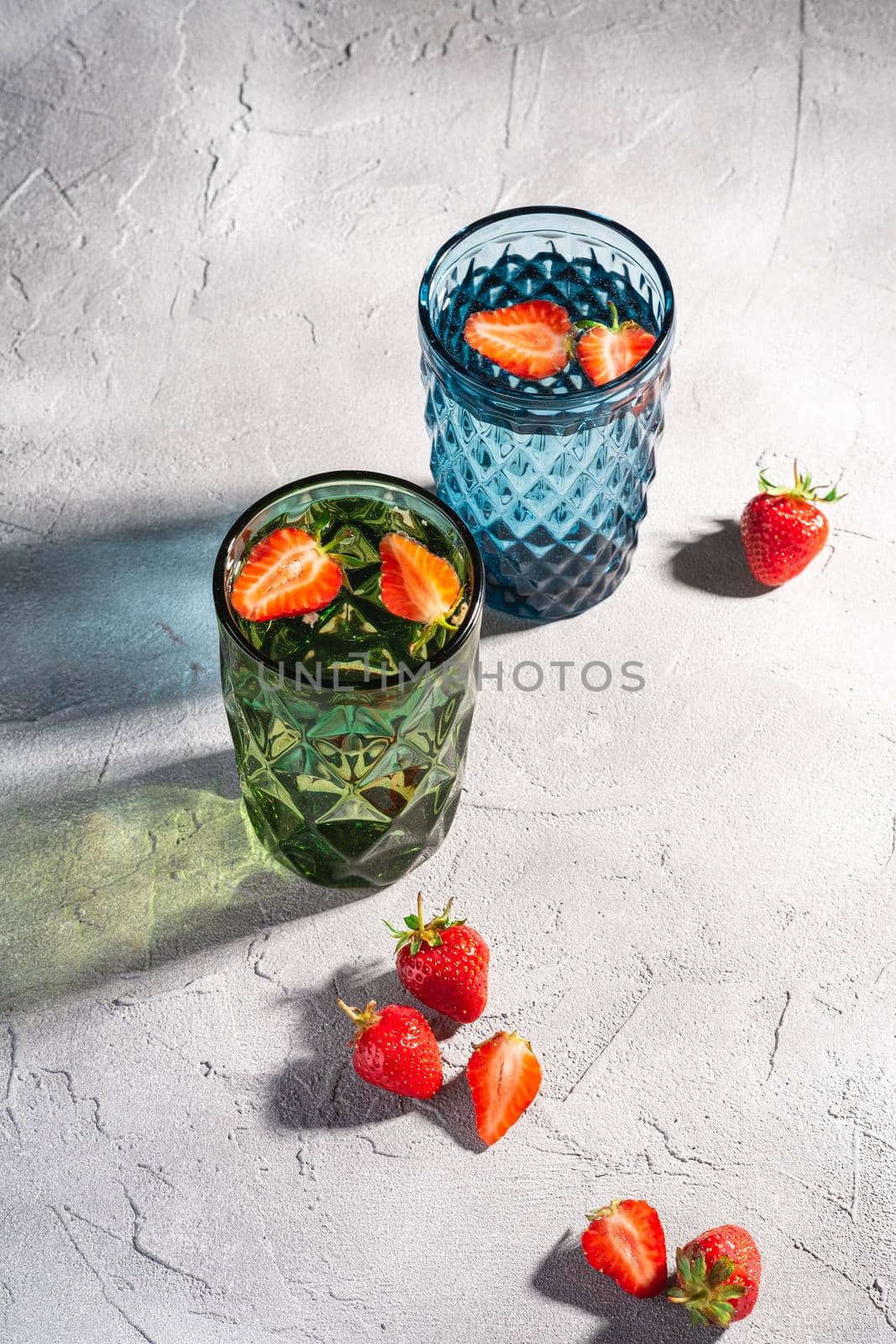 Two green and blue geometric glass cup with fresh water and strawberry fruits with colorful shadow light rays on stone concrete background, angle view