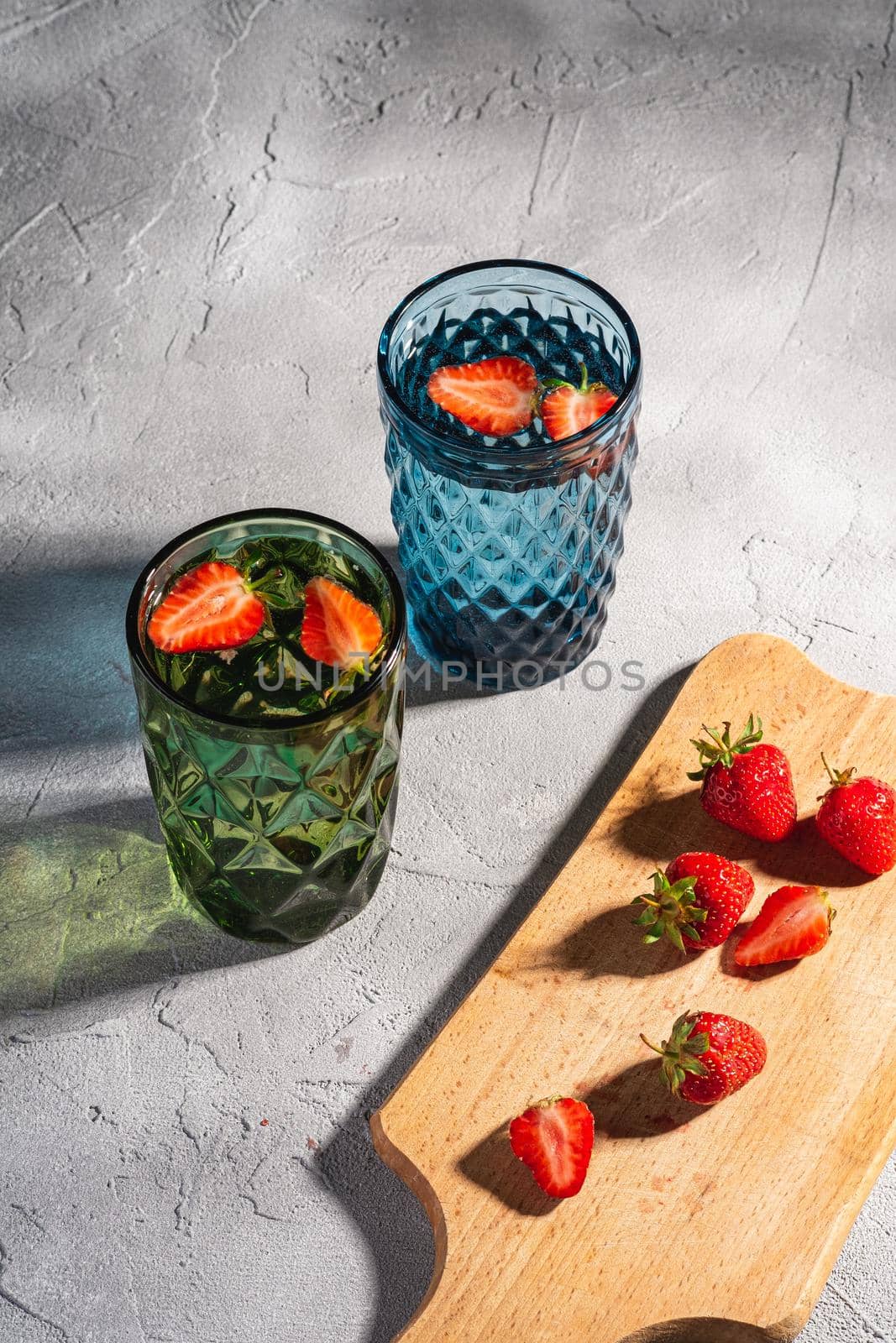 Two green and blue geometric glass cup with fresh water and strawberry fruits with colorful shadow light rays near to wooden cutting board on stone concrete background, angle view