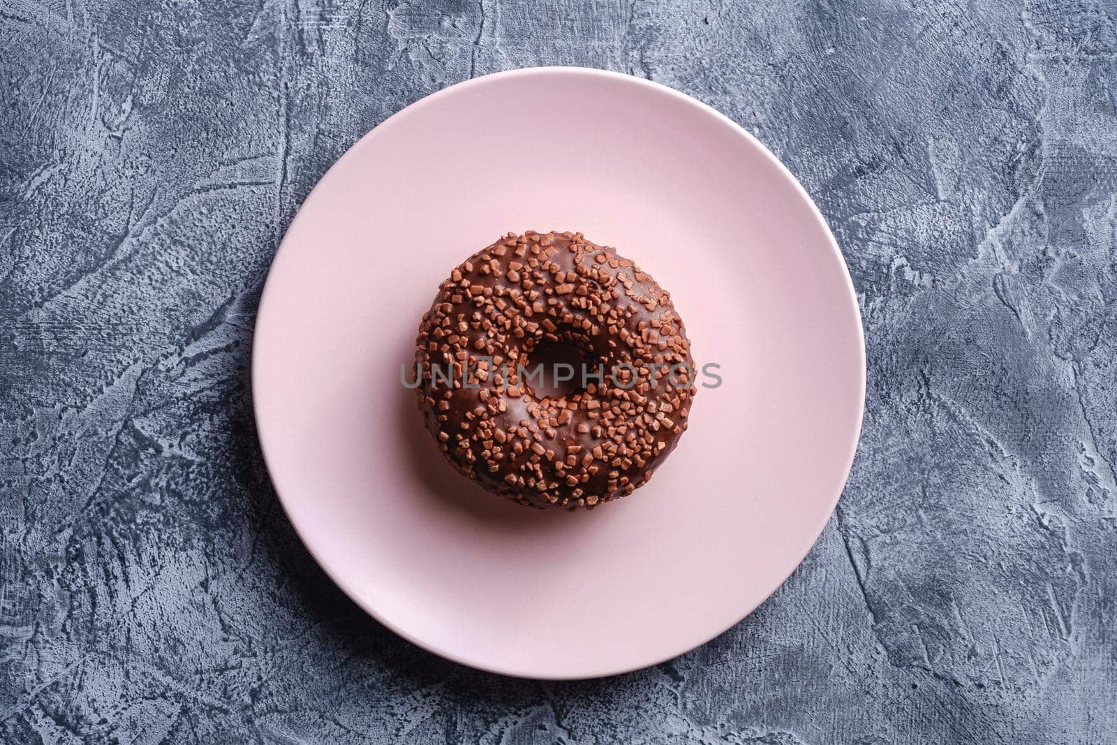 Chocolate donut with sprinkles on pink plate, sweet glazed dessert food on concrete textured background, top view