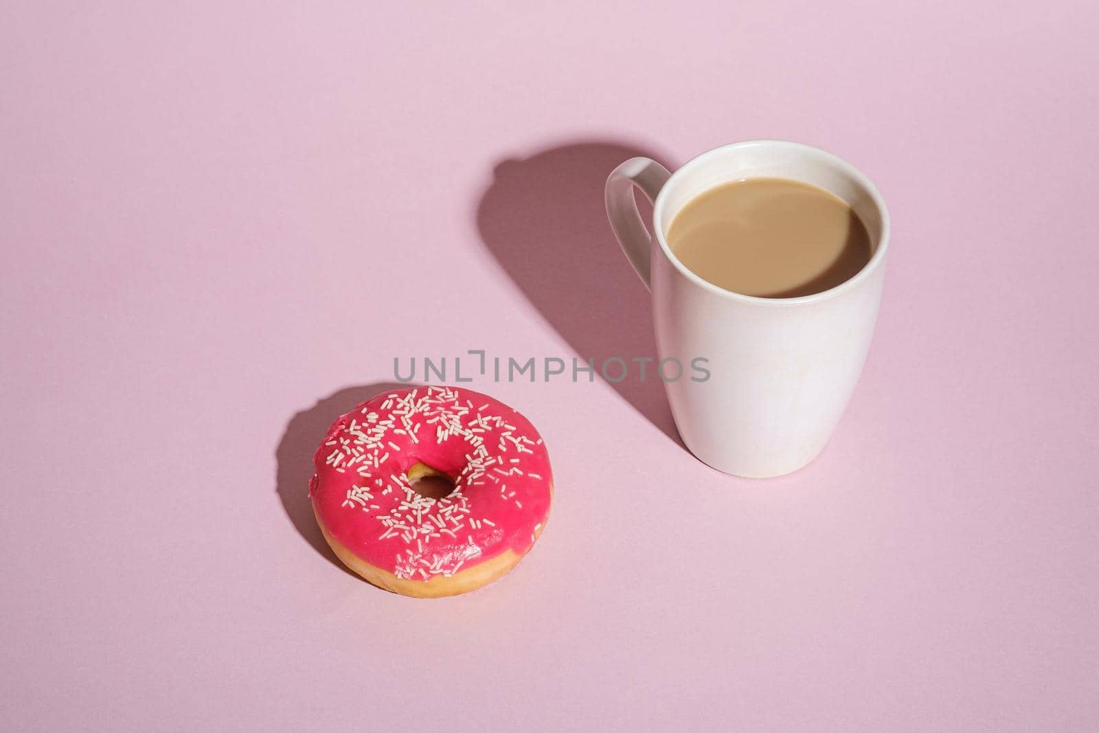 Pink donut with sprinkles near to cup of coffee, sweet glazed dessert food and hot drink on pink minimal background, angle view copy space