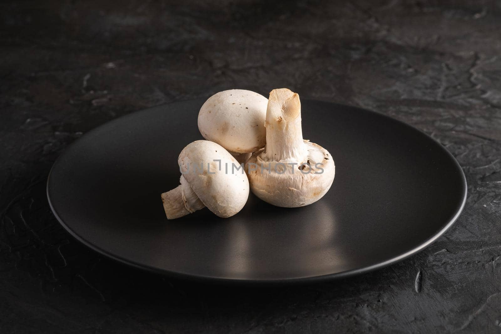 Three champignon mushrooms healthy food on black plate on dark black textured background, angle view macro