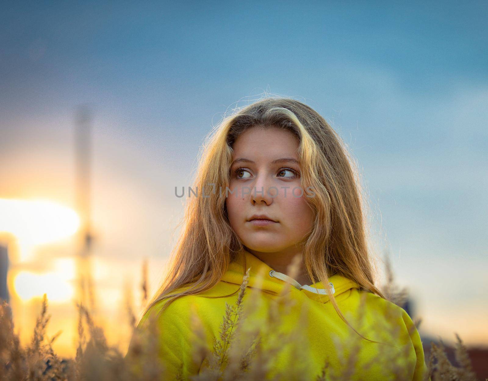 A young girl with long hair walks across a field with tall grass. Blonde hair. Yellow jacket.