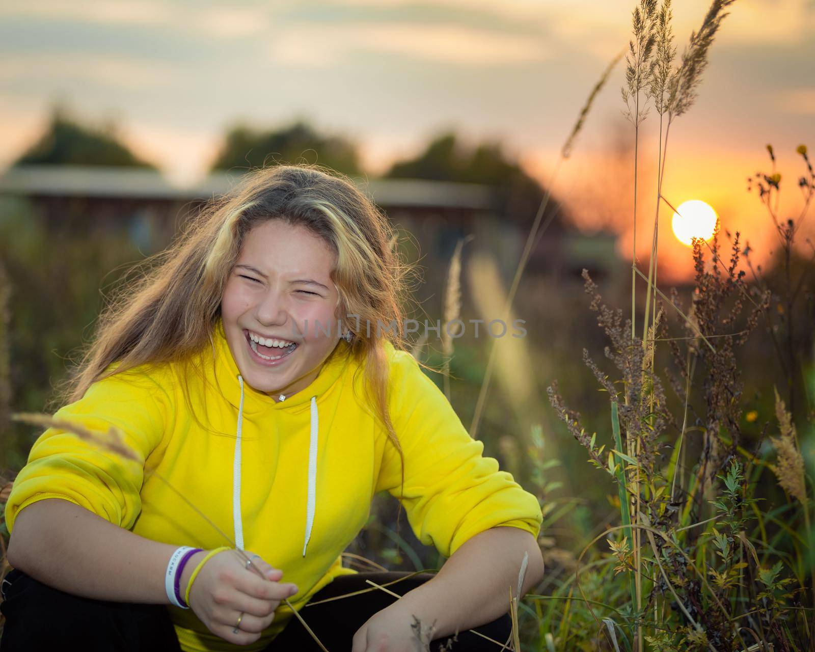 A young girl with long hair on a meadow with tall grass in the rays of the setting sun. Blonde hair. Yellow jacket.