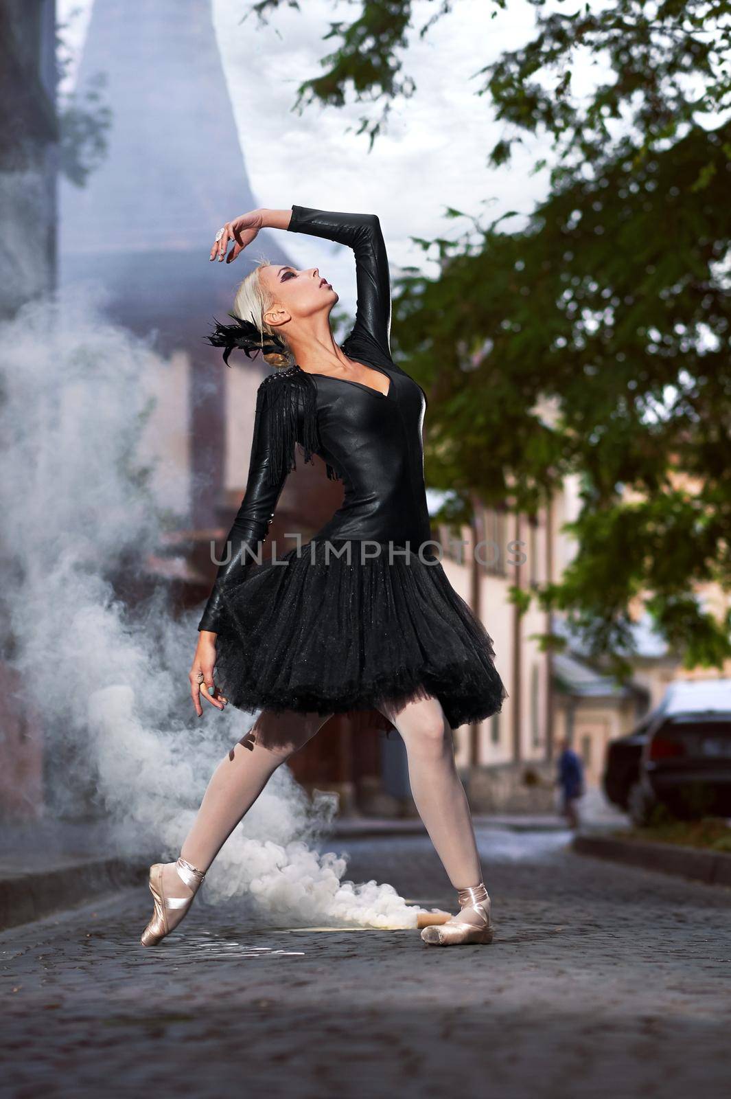 Vertical shot of a gorgeous professional female ballet dancer wearing black swan dancing outfit performing outdoors on the street of the city smoke on the background.