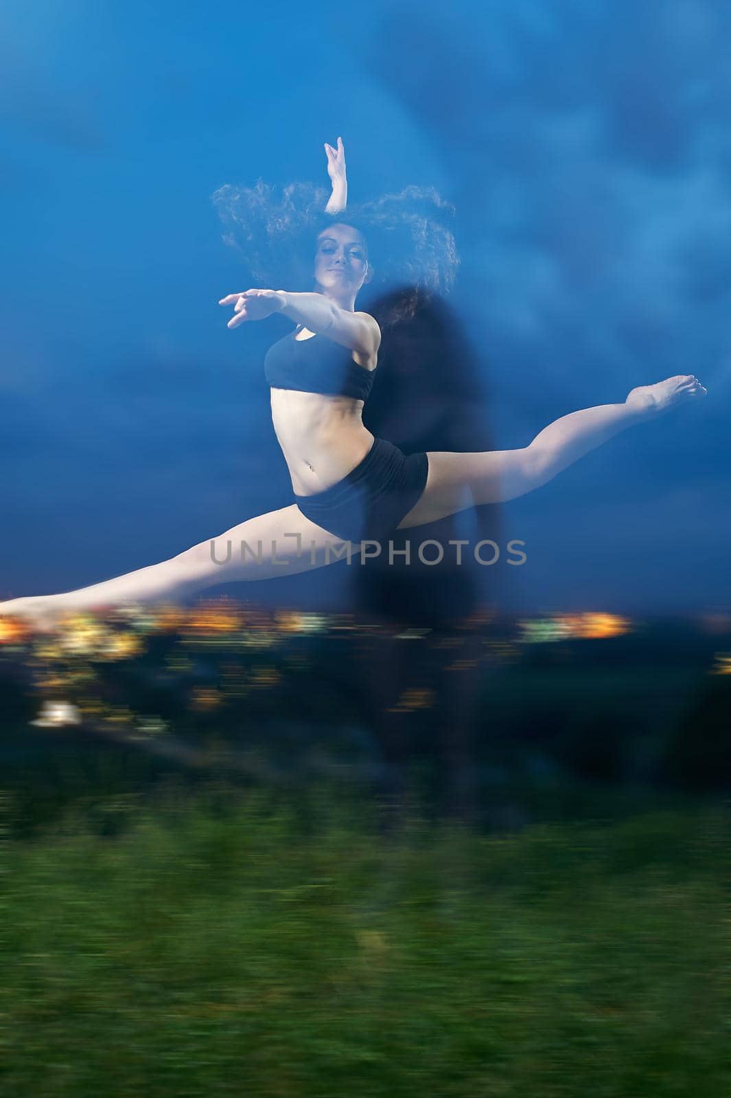 pretty, young, smiling dancer performs splits in the air at the grassy field in front of the night lights of the city.