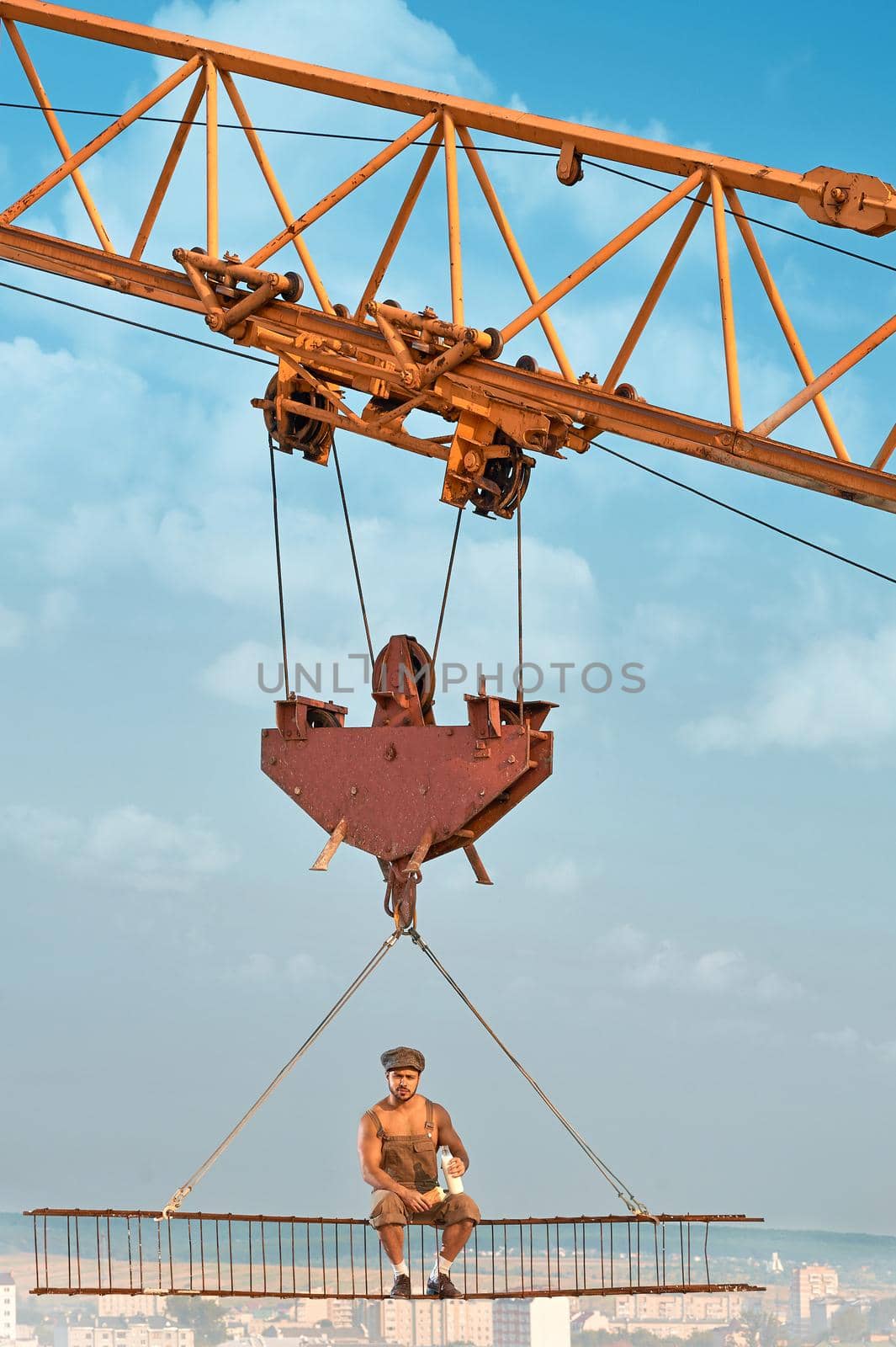 View from distance of bold builder in work wear sitting on construction on high and eating. Building crane holding construction with man over city. Extreme building of house in big city.