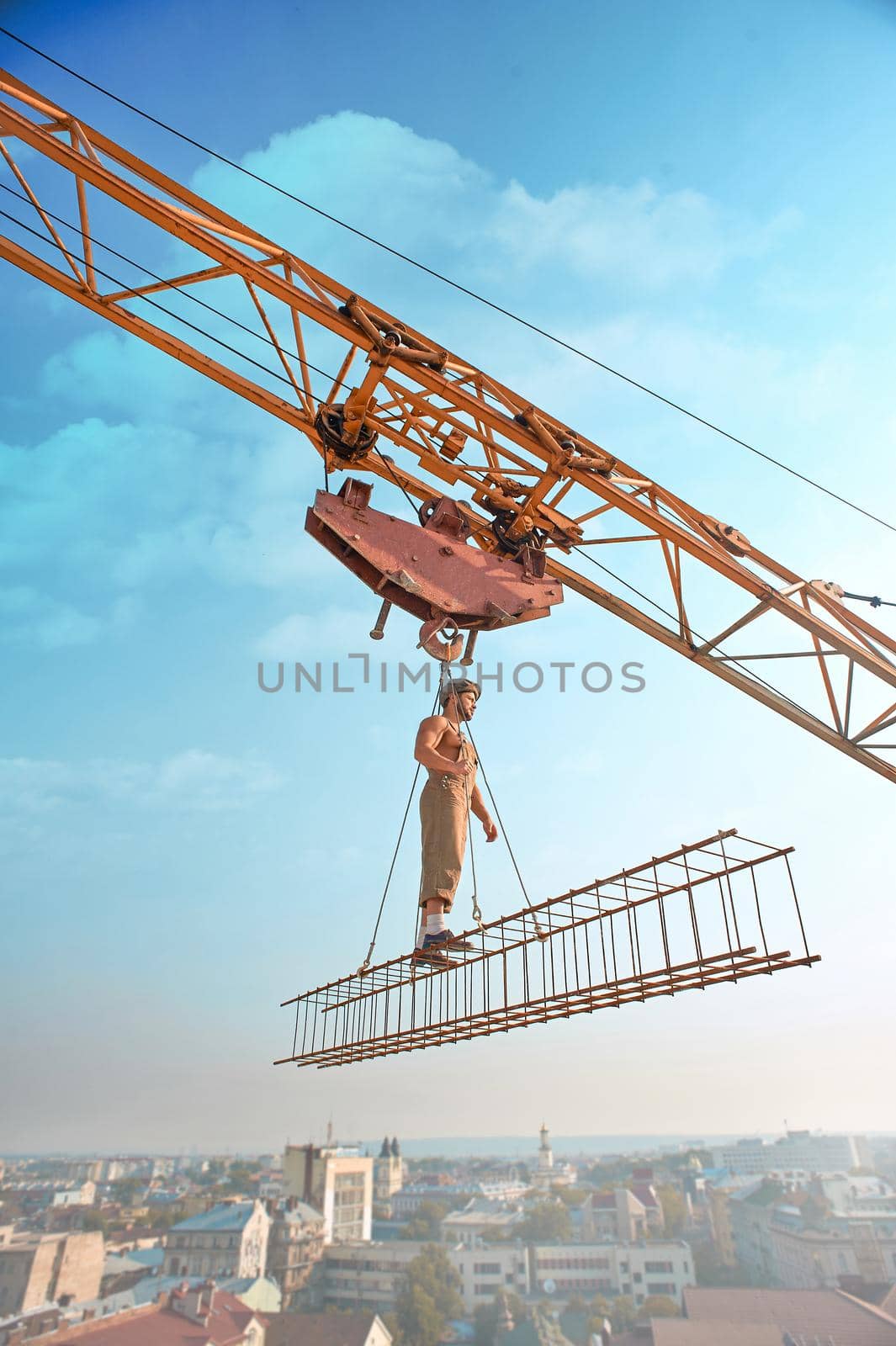 Side view of athletic man in hat standing on construction on high and looking away. Large building crane holding construction with male over city in air. Cityscape and blue sky on background.