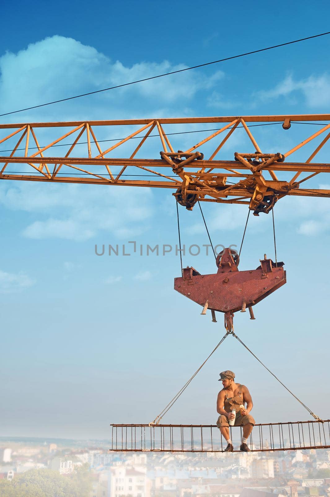 Front view of crane holding iron construction, where sitting muscular builder drinking milk and eating bread. Extreme building on high over city. Blue sky on background.