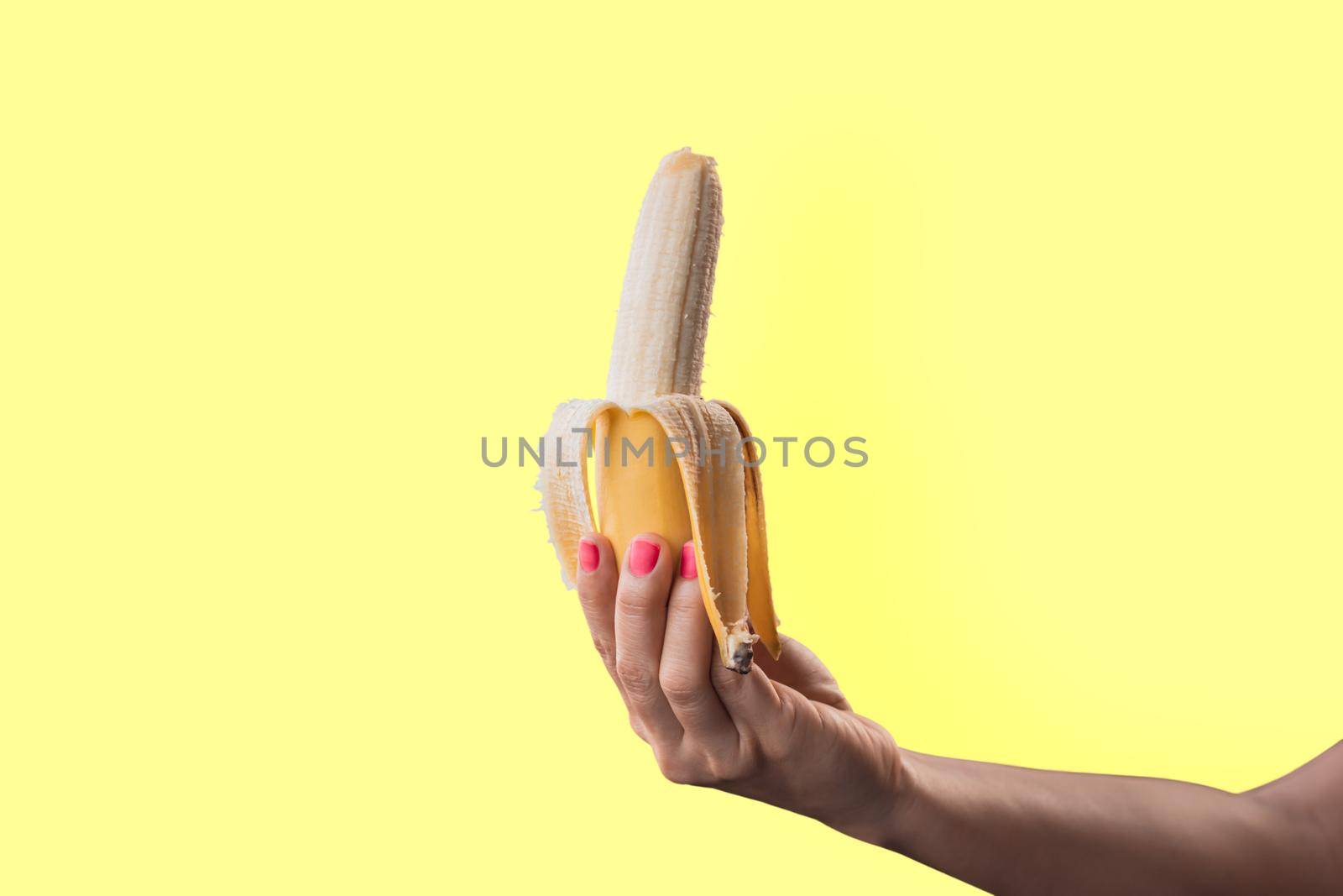 Closeup a woman's hand painted red nails holding a ripe banana isolated on yellow pastel colors background by wattanaphob