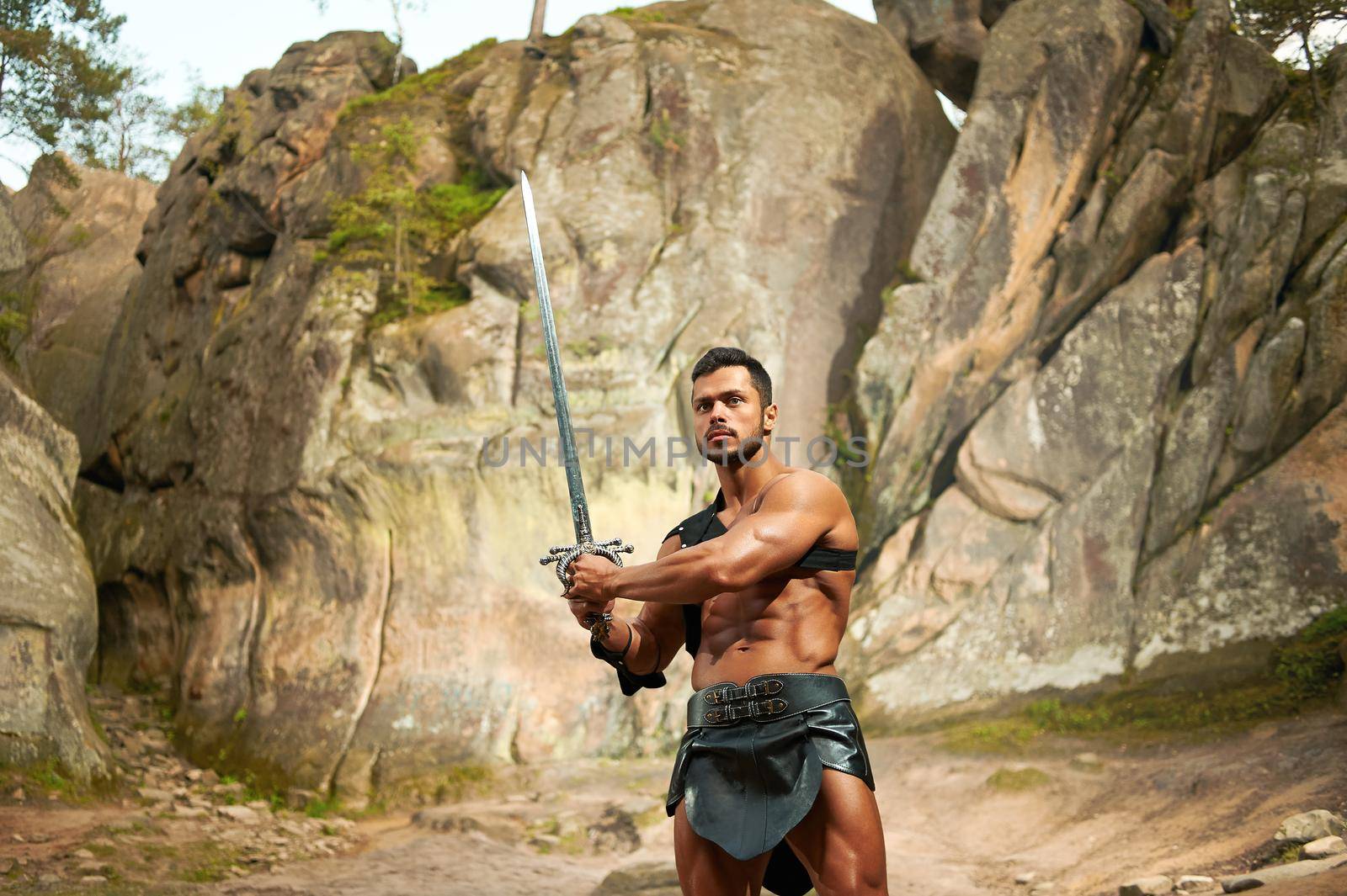 Not afraid to face his enemies. Brave young warrior with strong muscular body holding his sword standing near the rocks