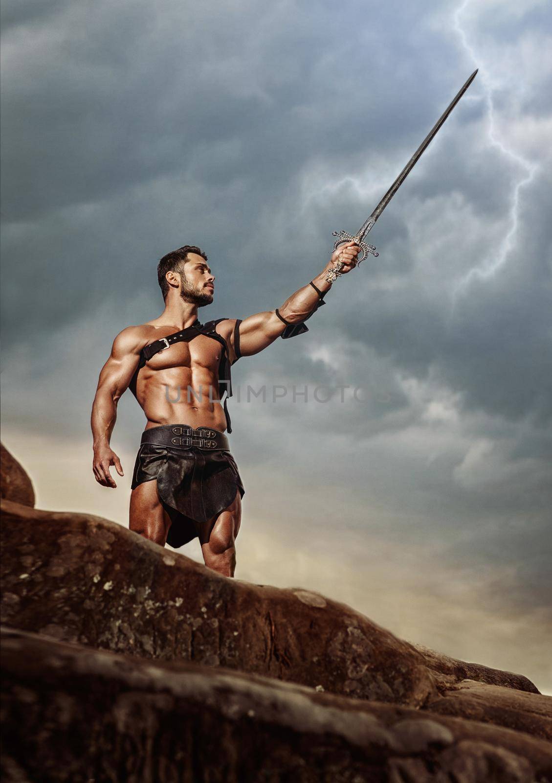 Proud warrior. Vertical low angle shot of a strong muscular Spartan warrior pointing his sword at the grey stormy skies