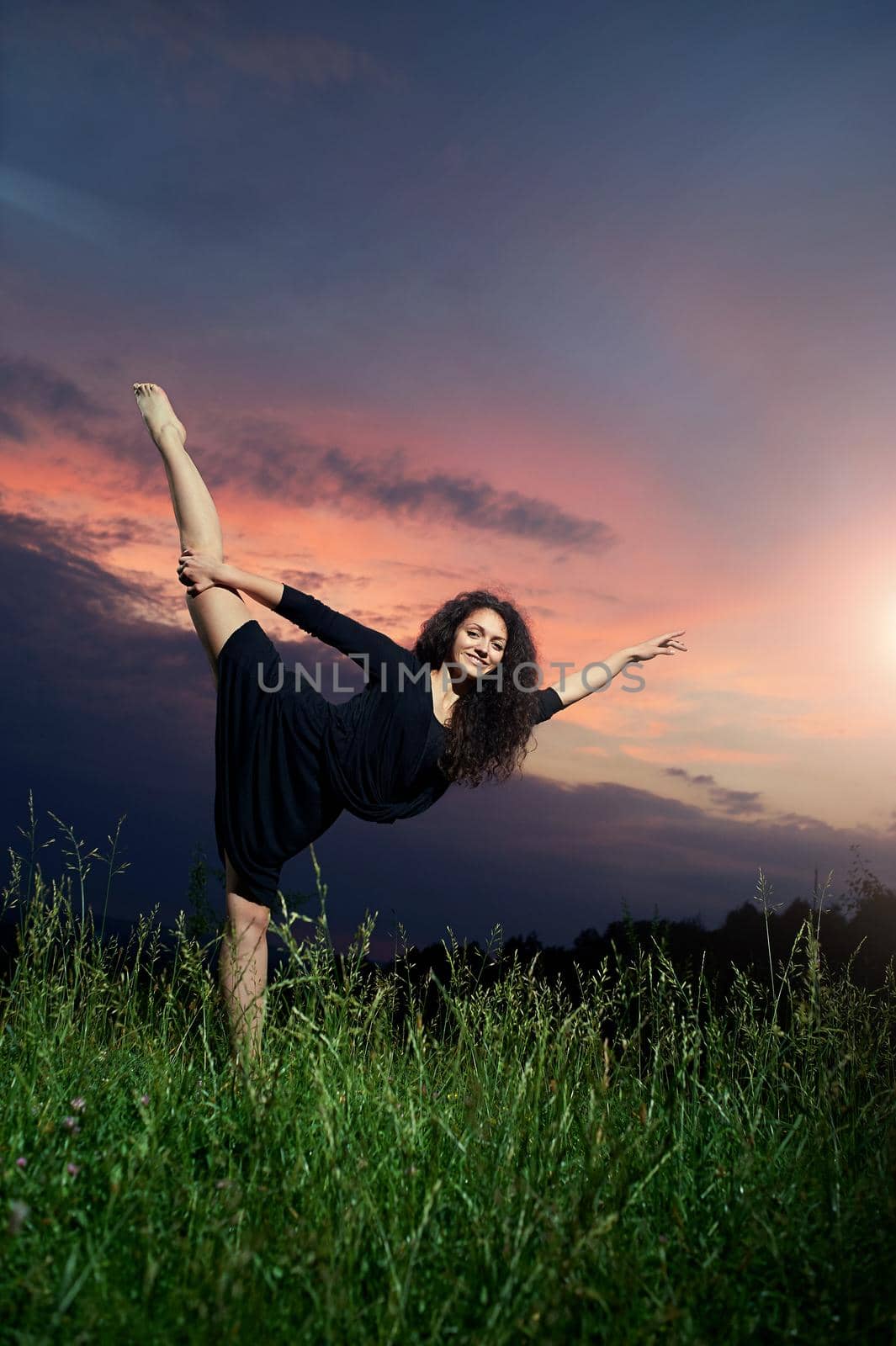 beautiful, flexible, athletic, curly dancer performs vertical side splits in grassy field at sunrise.