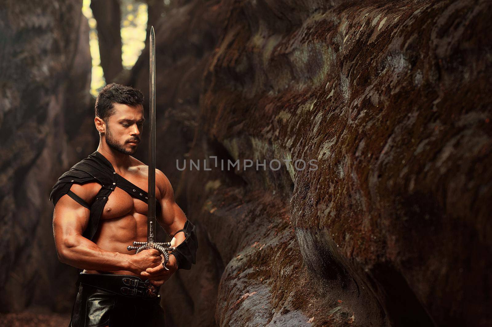 Deep in his thoughts. Horizontal portrait of well-built muscular young warrior standing with a sword with his eyes closed near the rock at the woods