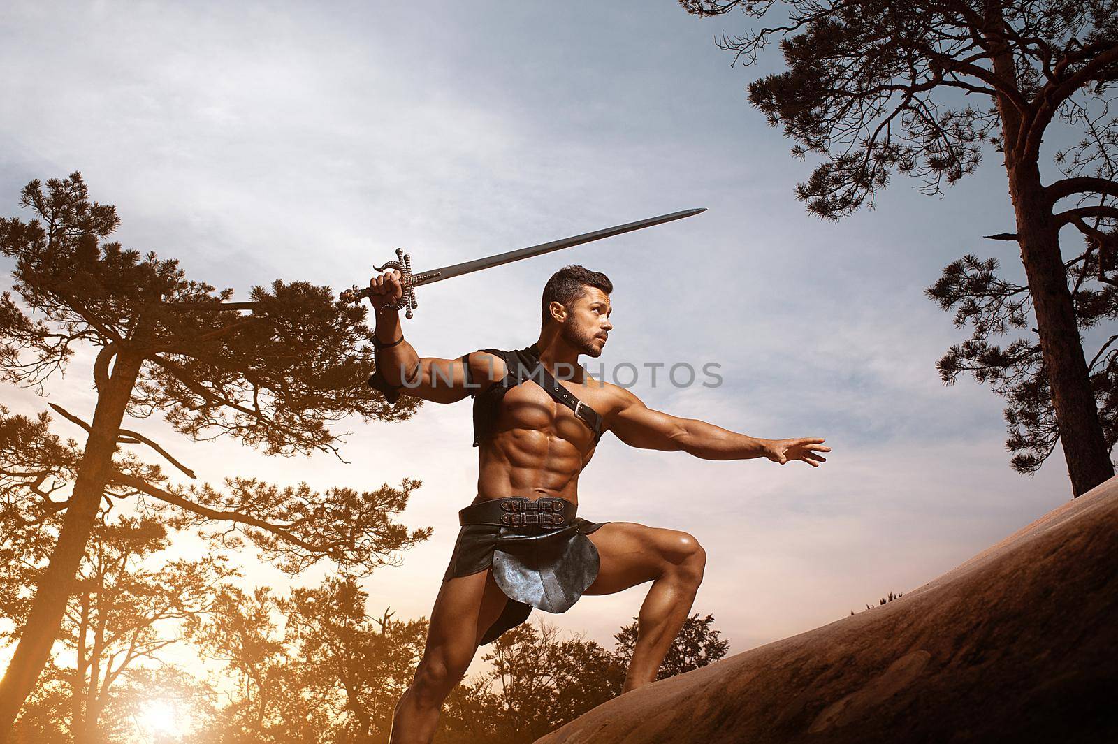 Graceful masculinity. Young muscular warrior with a sword lurking carefully in the woods at the dusk copyspace