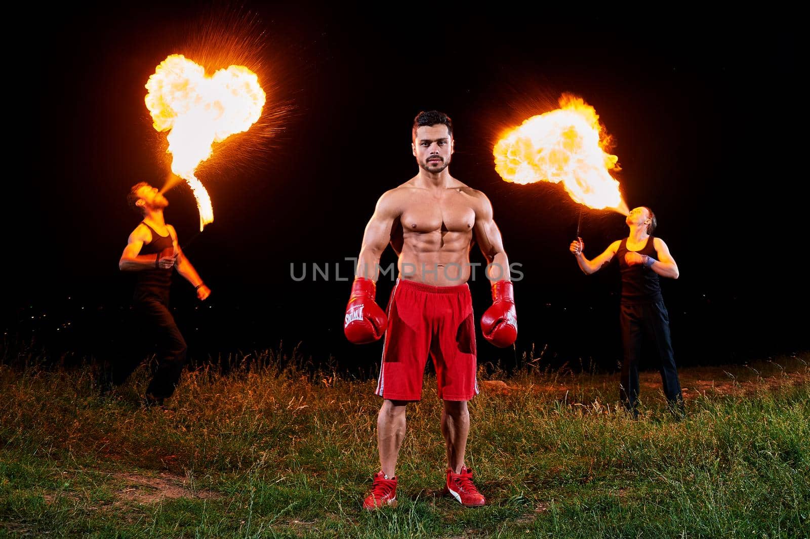 Handsome young muscular shirtless boxer man wearing boxer gloves posing outdoors extreme fire show on the background people blowing fire from their mouths copyspace exotic unusual sport sexy hot.