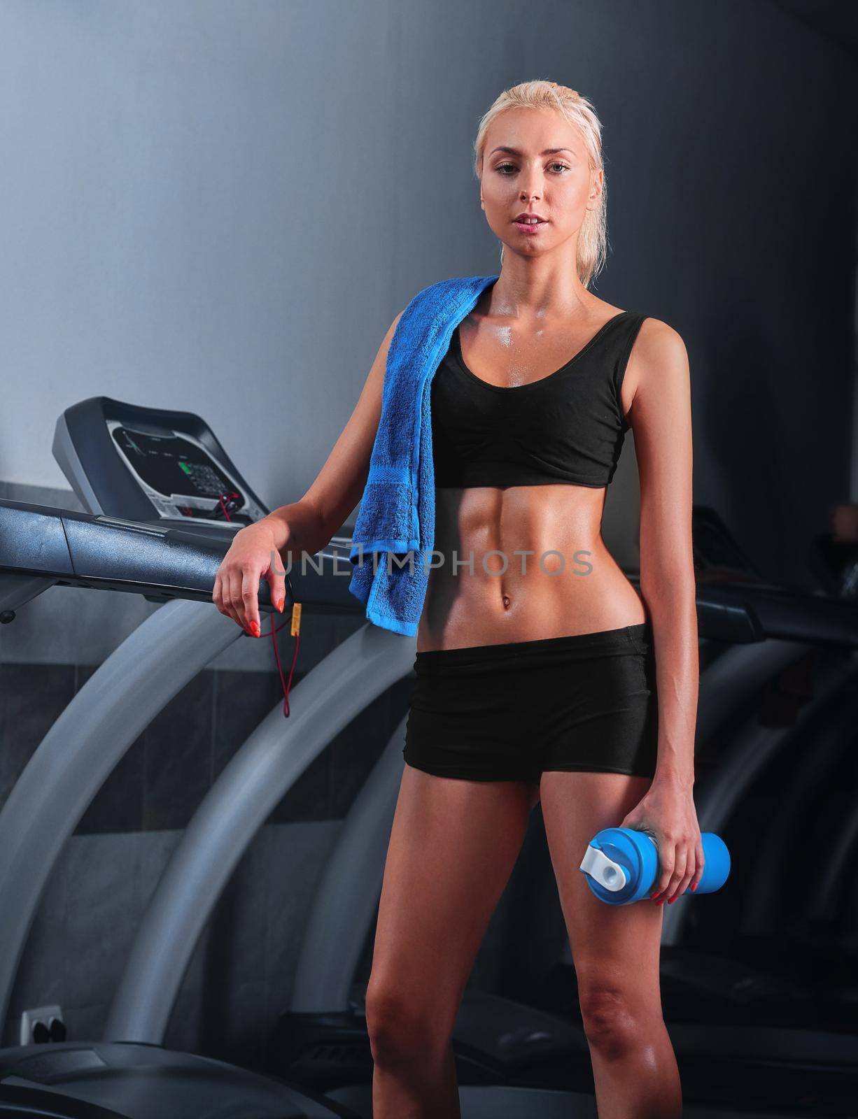 Vertical portrait of a gorgeous blonde fitness woman smiling to the camera standing near treadmill after her workout at the gym resting relaxing lifestyle happiness towel water body strong abs muscles.