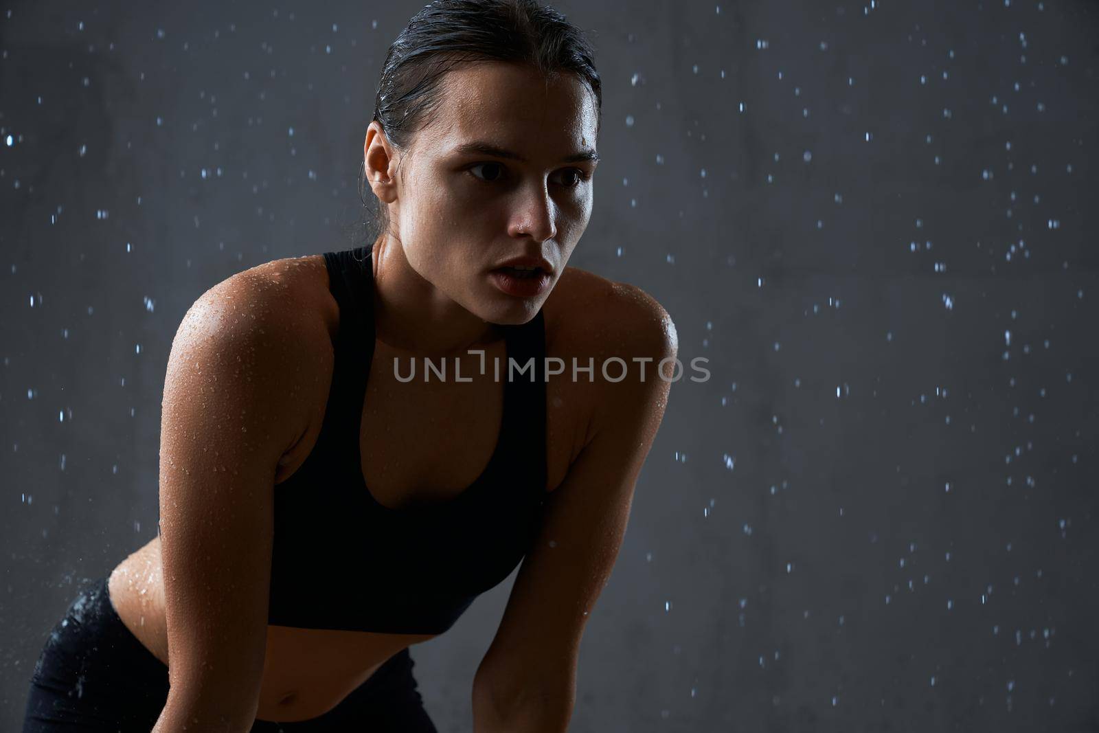 Close up of muscular fitnesswoman posing under rain. Crop of wet fit female model holding hands on knees, wearing black sportswear having rest after hard training, isolated on gray.