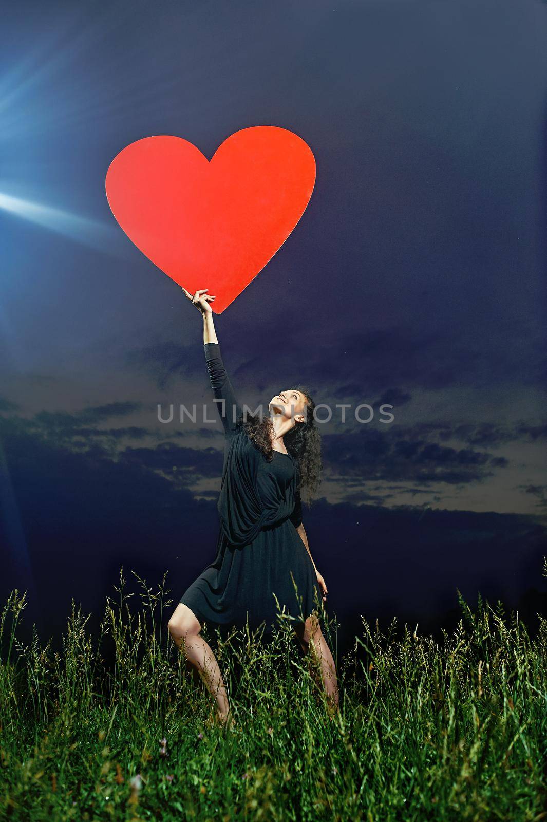 beautiful, flexible, smiling, curly dancer posing on grassy field with a red heart in his hands in front of the evening sky.