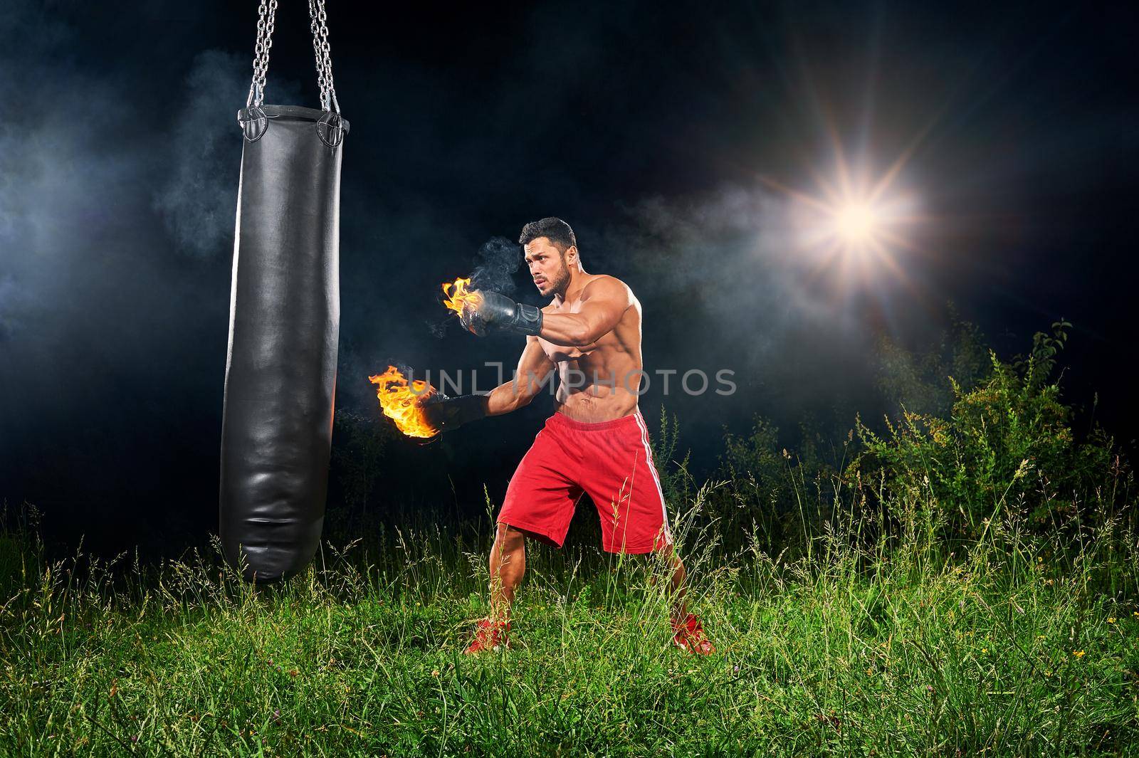 Young athletic male fighter working out with a punching bag outdoors at night with fire burning on his boxing gloves copyspace power strength masculinity fierce fiery flames hot sexy sportsman focused.