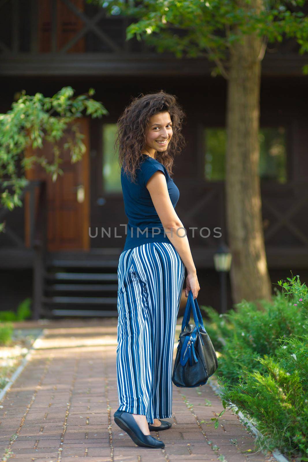young beautiful brunette with curly hair, in a stylish outfit with a bag. Enjoying a bright summer day in a green park.