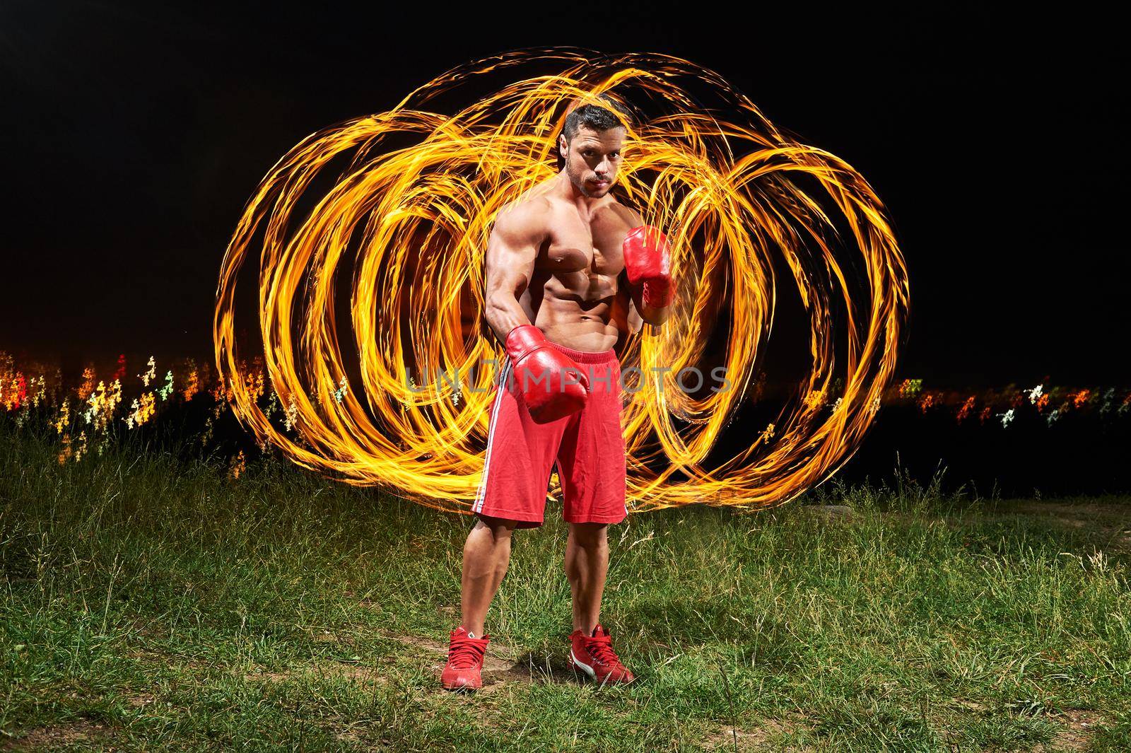 Muscular shirtless handsome sexy young man wearing boxing gloves posing forcefully outdoors at night with fire and flames behind his back copyspace sportsman sport motivation athlete winner fighting .