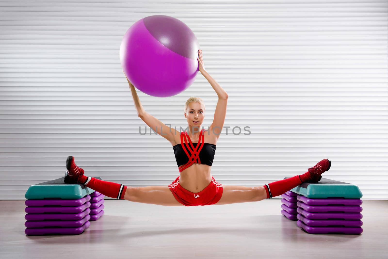 Stunning flexible female gymnast holding a fit ball over her head while practicing splits at the fitness studio flexibility stretching exercise workout trainer body strength motivation sports concept.
