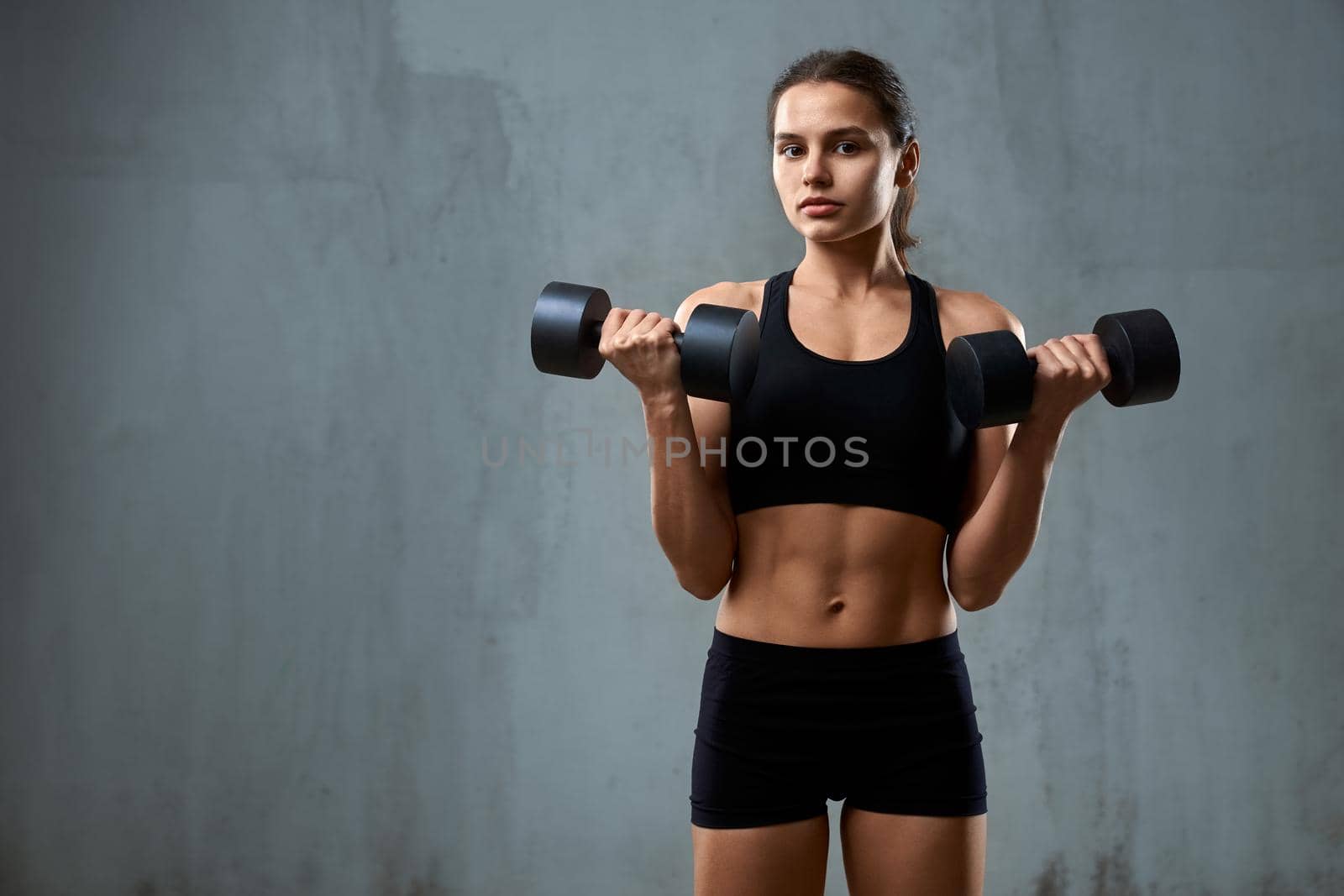 Isolated portrait of pumped fitnesswoman in black sportswear posing on gray studio background, loft interior. Front view of muscular caucasian woman training with dumbbells and looking at camera.