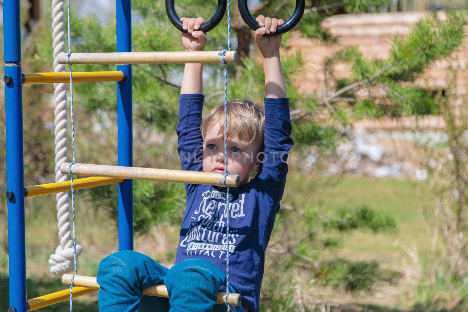 A blonde-haired child is playing on the playground, hanging on rings.
