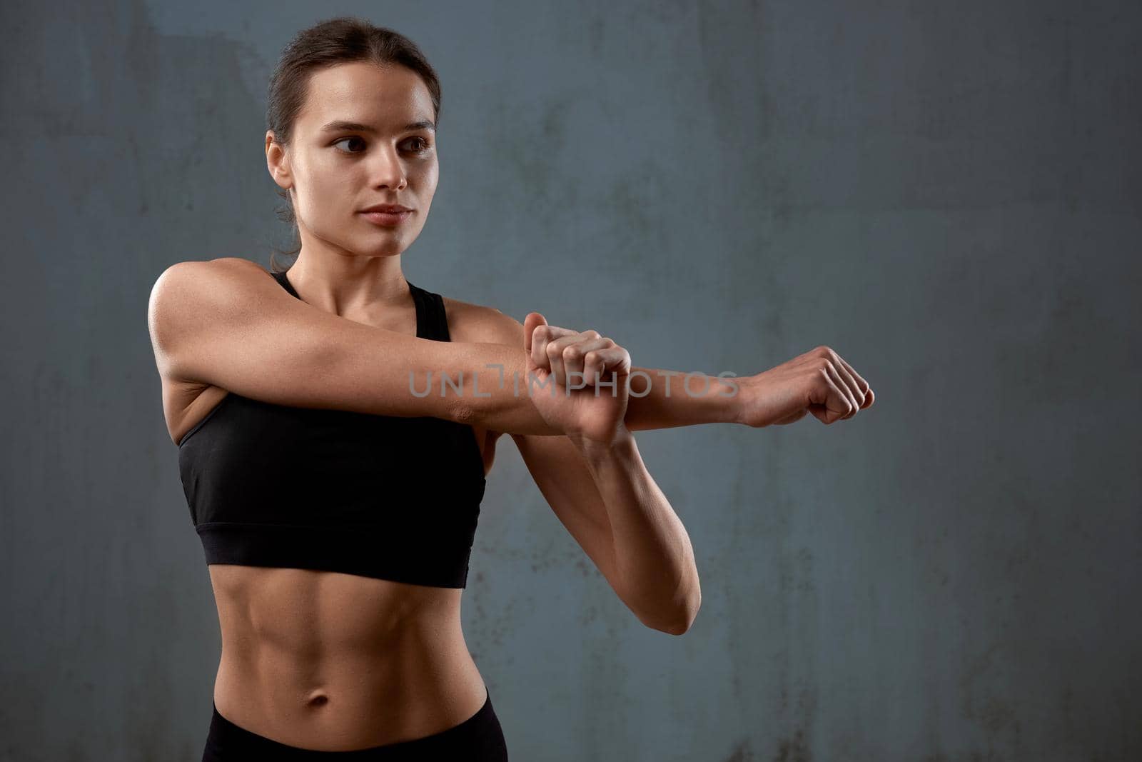 Close up portrait of fit female model stretching arm before training, isolated on loft gray studio background. Muscular flexible young fitnesswoman posing in black sports underwear indoor.