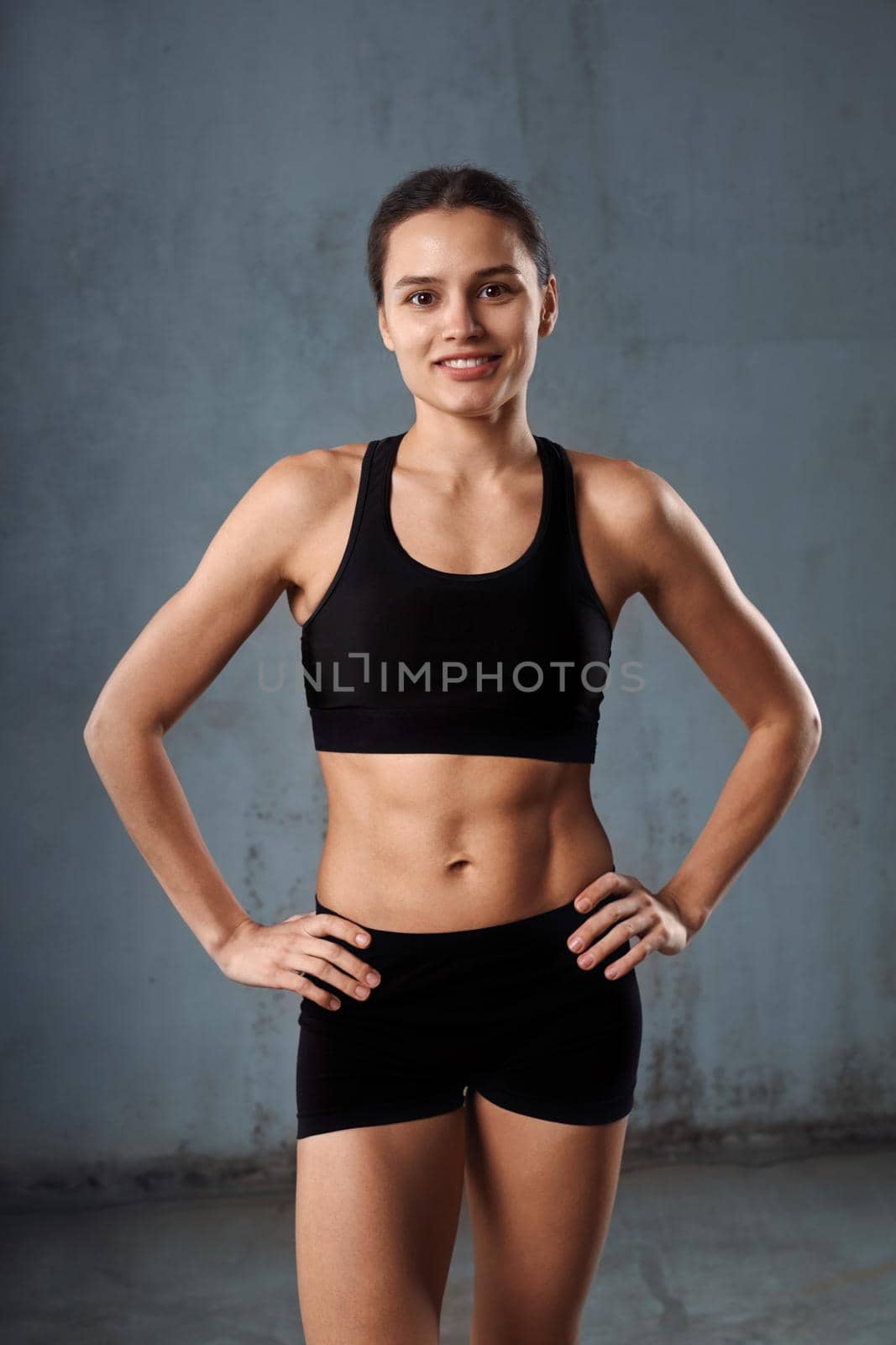 Portrait of smiling fit female model holding hands on waist, wearing black sportswear standing, isolated in loft gray studio. Stylish pretty young fitnesswoman with ponytail looking at camera.