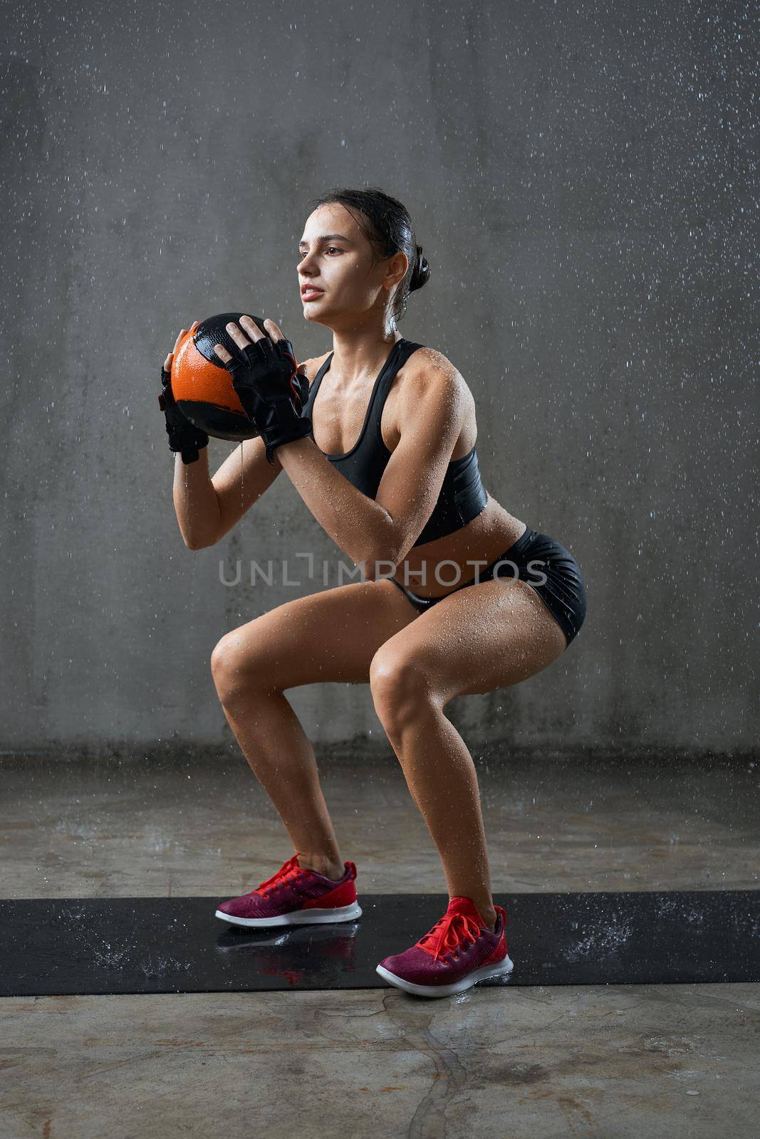 Side view of pretty female bodybuilder in gloves training legs, holding small ball in hands under rain. Wet muscular sportswoman with perfect body doing squats on mat in empty hall, loft interior.