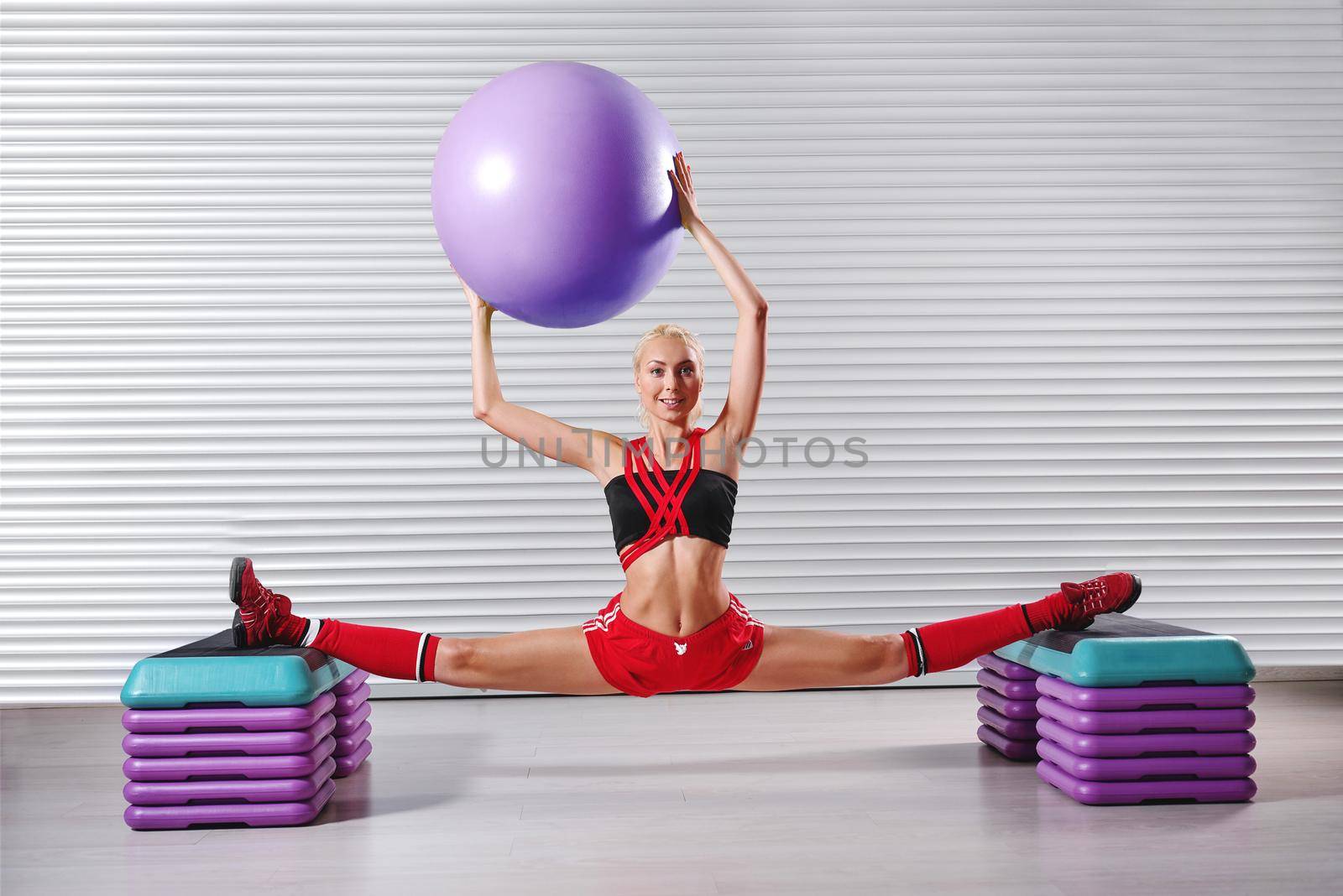 Horizontal shot of a happy female gymnast doing splits at her fitness studio smiling to the camera holding a fit ball over her head sports flexibility stretching body strength confidence gymnastics .