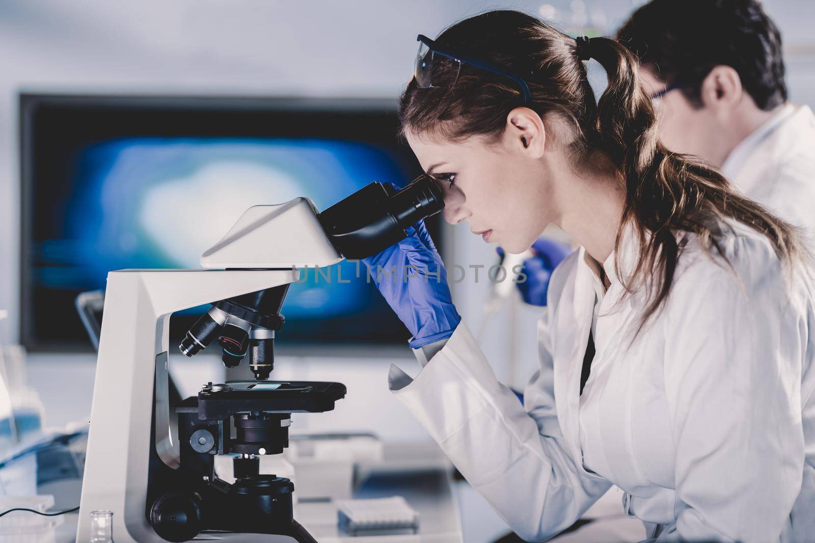 Life scientists researching in laboratory. Attractive female young scientist and her post doctoral supervisor microscoping in their working environment. Healthcare and biotechnology.